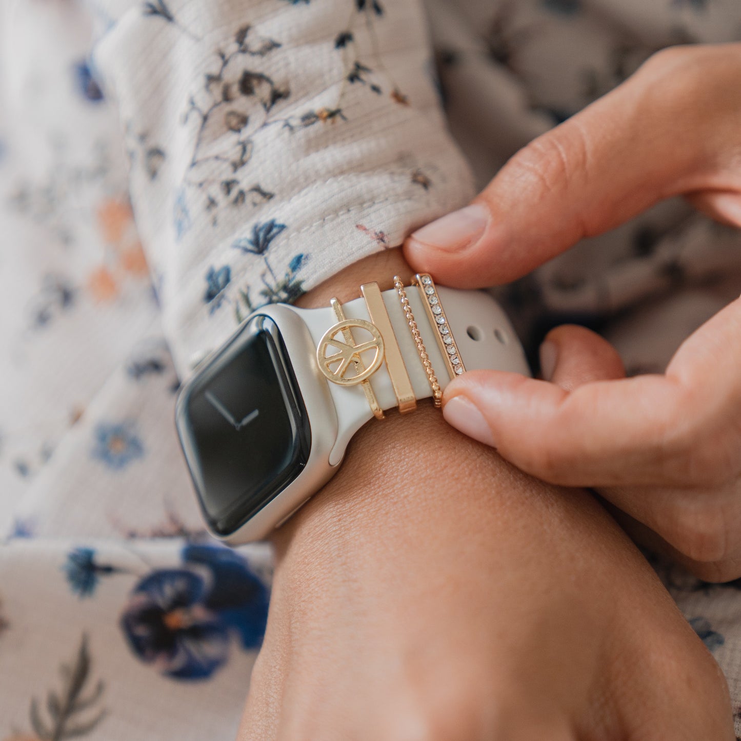 Eine Person in einem hellen Hemd mit Blumenmuster rückt ihr weißes Apple Watch-Armband zurecht, das mit arktisband Apple Watch-Armband-Anhängern in Gold- und Juwelendesigns von arktisband verziert ist.