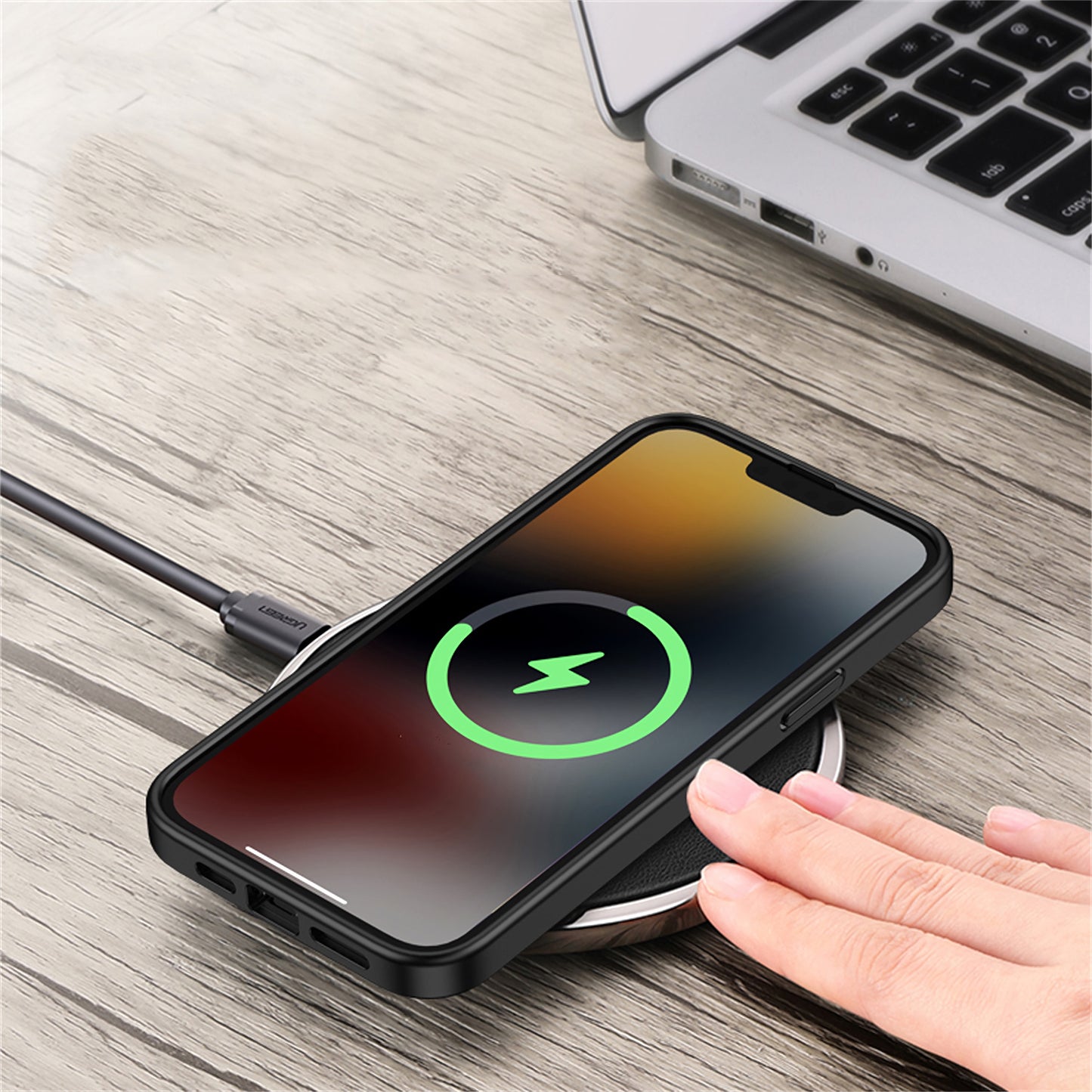 Eine Hand legt ein iPhone 13 mit dem Aviato SECTOR SLIM Case – Full Black in einer kabellosen Ladestation neben einem offenen Laptop auf einer Holzoberfläche.