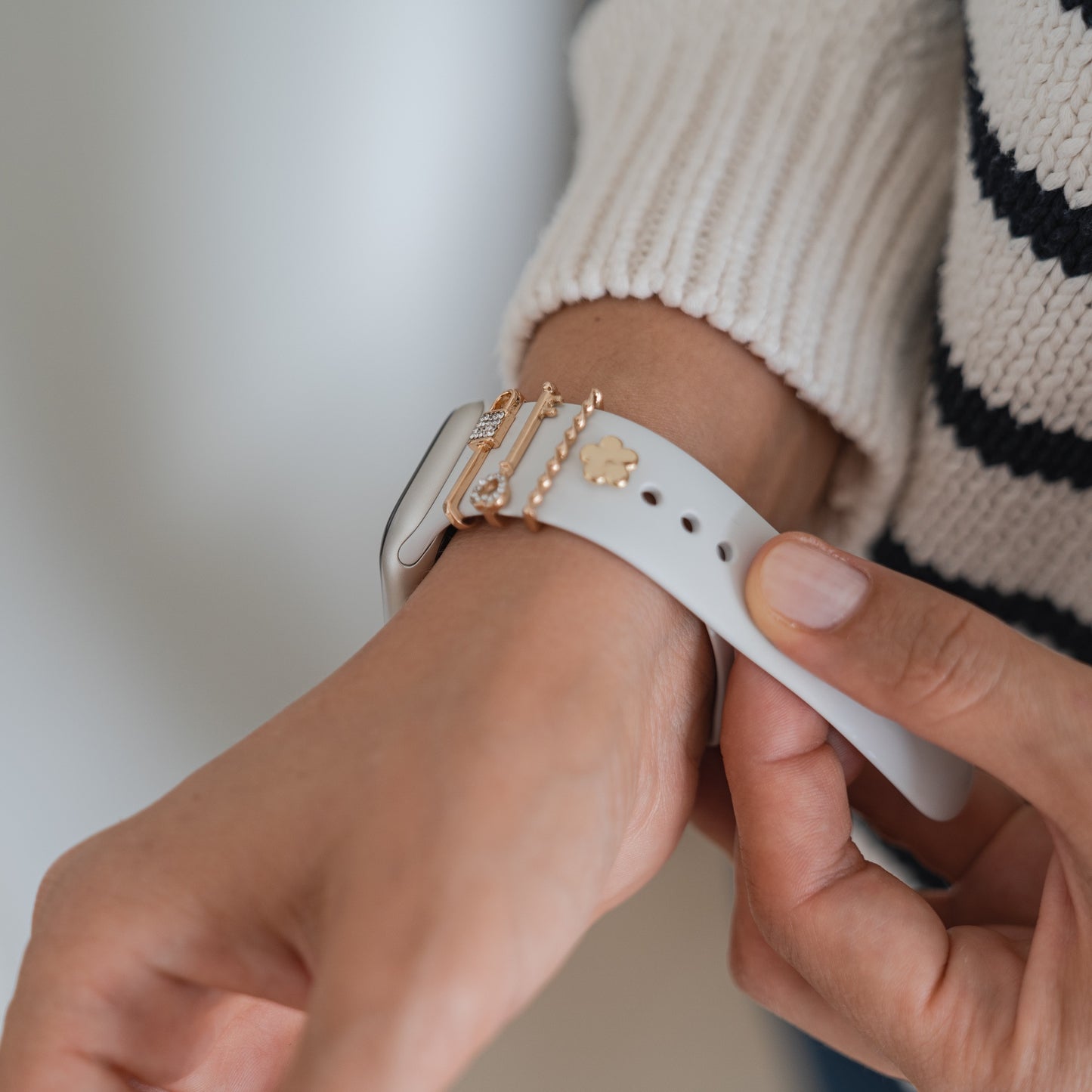 Eine Person trägt einen cremefarbenen Pullover mit schwarzen Streifen und befestigt ein arktisband Apple Watch Armband Charm – ein weißes Smartwatch-Armband mit einem goldenen Charm von arktisband – an ihrem Handgelenk. Sie zeigt, wie sich der Look individuell gestalten lässt. Nur ihre Hand und ihr Arm sind sichtbar.