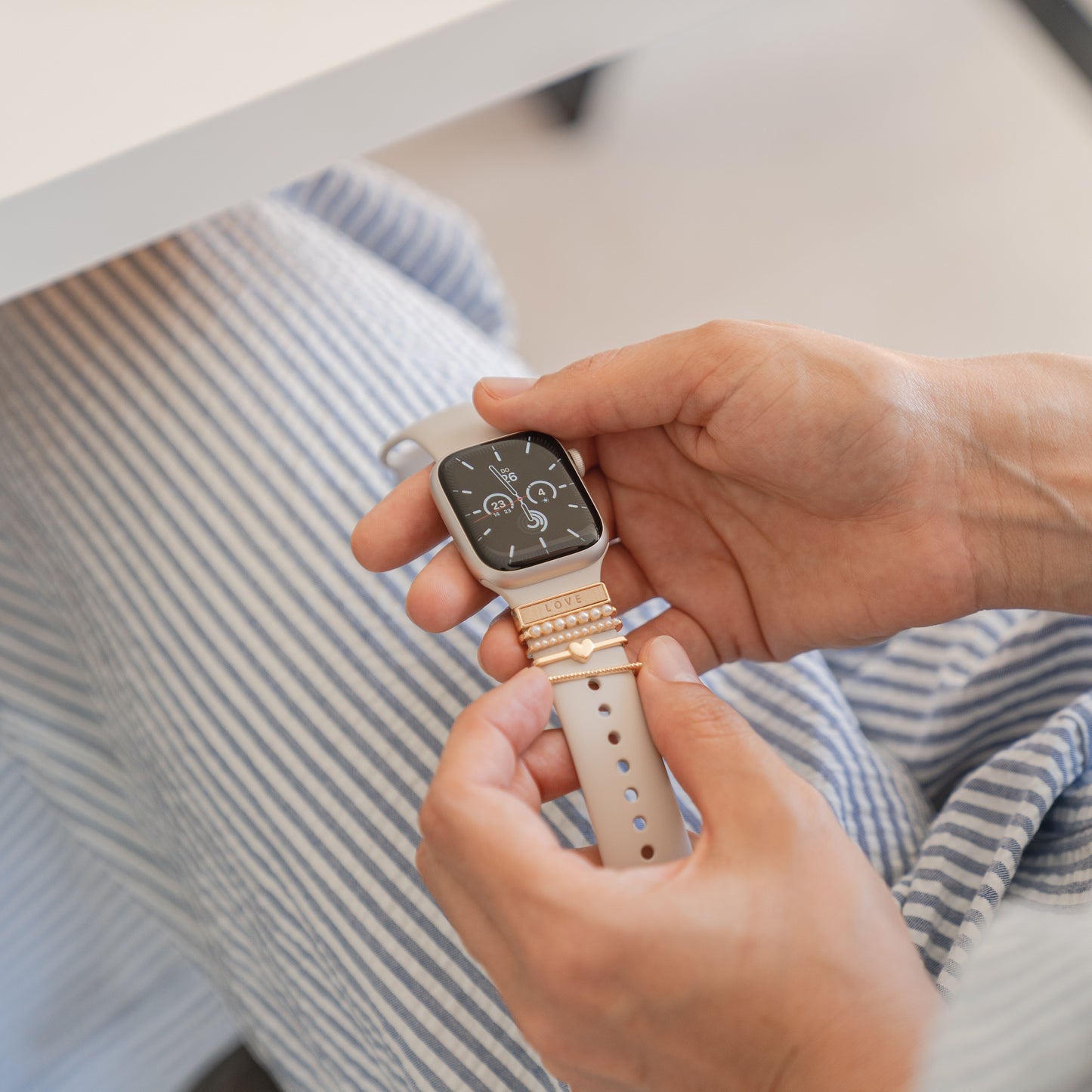 Eine Person in gestreiften Hosen hält eine Apple Watch mit einem beigen Arktisband und Arktisband Apple Watch Armband-Anhängern neben einem weißen Tisch. Der Bildschirm der Smartwatch zeigt verschiedene digitale Zifferblätter und Informationen an.