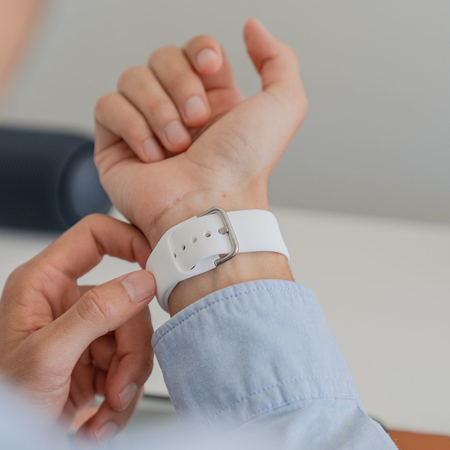 Eine Person im hellblauen Langarmshirt befestigt ein arktisband Apple Watch Silikonarmband „Aqua“ von arktisband um ihr linkes Handgelenk, vor einem sanft verschwommenen, hellen Hintergrund.