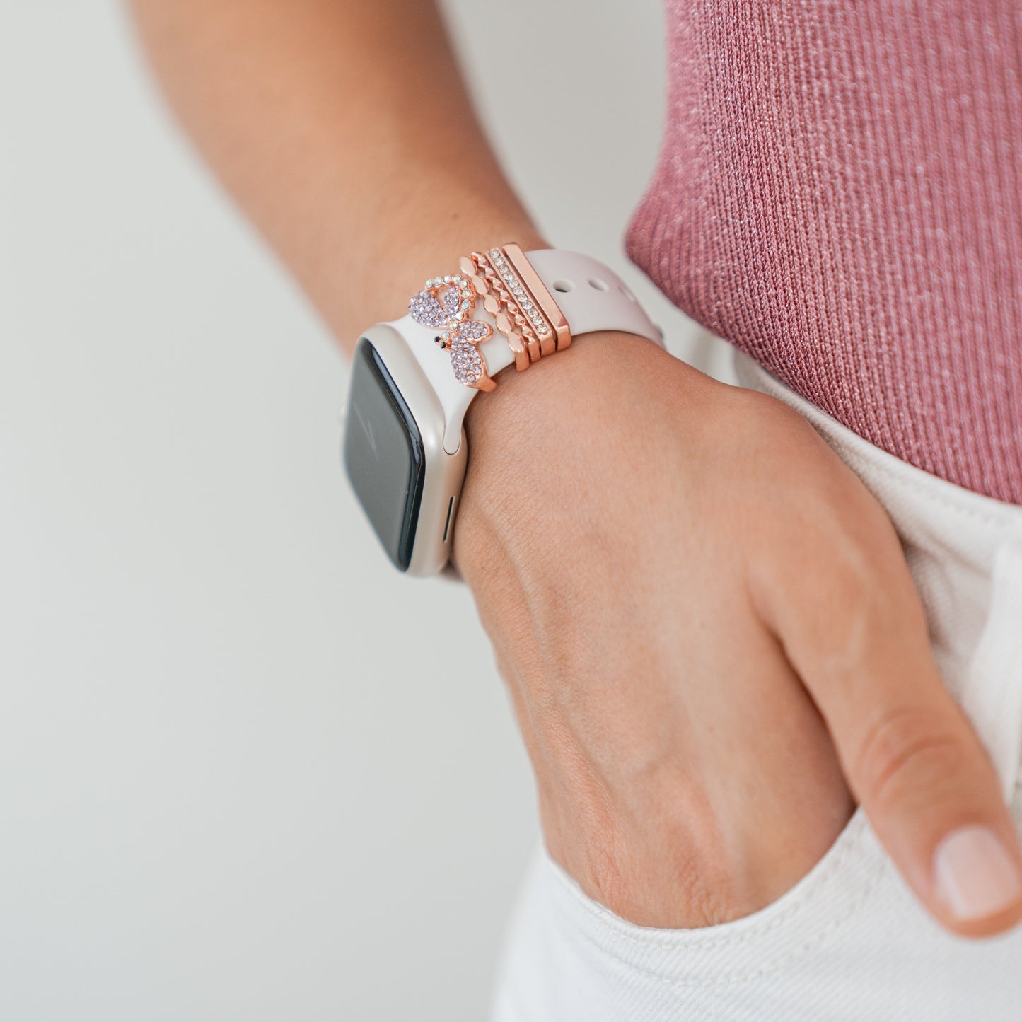 Sie tragen ein arktisband Apple Watch Armband Charms von arktisband mit Roségold- und Kristall-Akzenten und stecken eine Hand in die Tasche einer weißen Hose. Ein rosa geripptes Oberteil rundet den Look ab.
