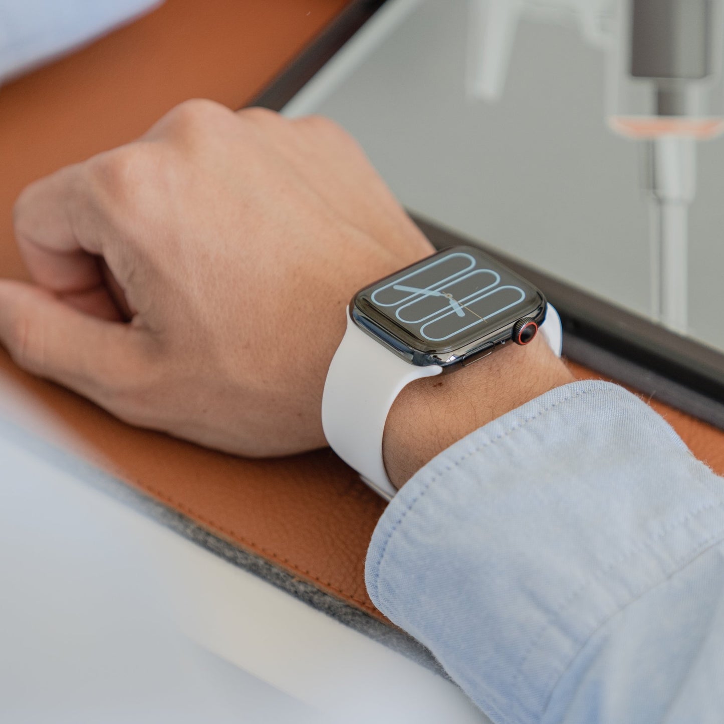 Eine Person trägt ein Arktisband Apple Watch Silikonarmband „Aqua“ und eine weiße Apple Watch kompatible Smartwatch am linken Handgelenk. Ihr Arm liegt auf einer braun-grauen Fläche neben einem Tablet. Das Uhrendisplay zeigt weiße digitale Zahlen und Linien.