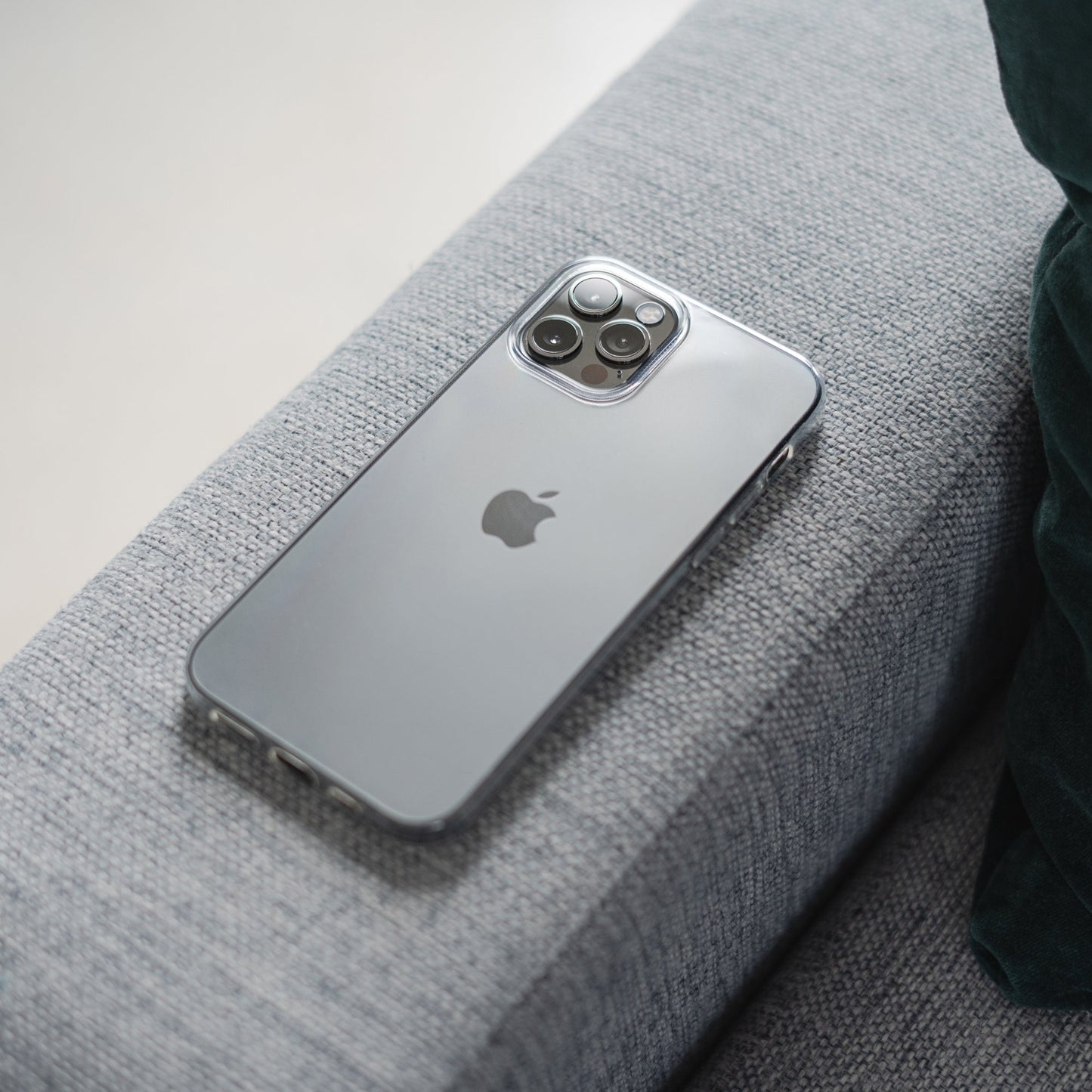 Ein ArktisPRO iPhone 14 Pro Max mit dem ZERO Case Ultra Clear liegt mit der Vorderseite nach unten auf der Armlehne eines hellgrauen Stoffsofas neben einem dunkelgrünen Kissen.