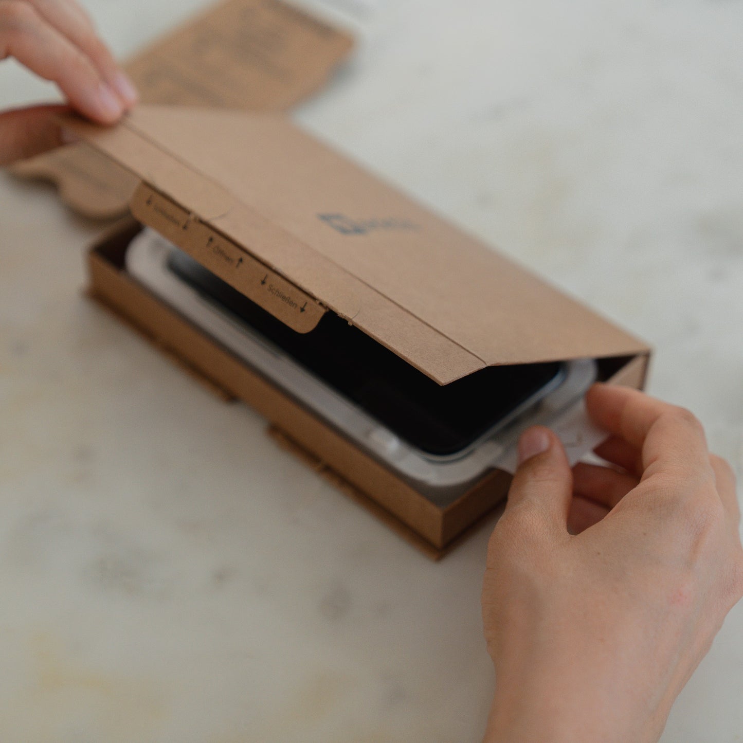 Hände, die eine EZ.INSTALL-Box mit einem schwarzen Smartphone und dem arktis 2er Set iPhone 17 Pro Max REVOLUTION Glas-Displayschutz auf einer weißen Fläche öffnen.