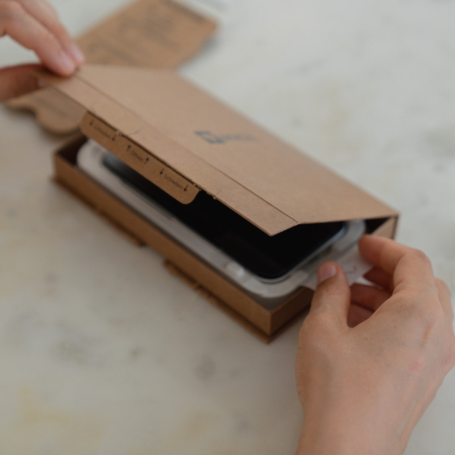 Hände, die eine EZ.INSTALL-Box mit einem schwarzen Smartphone und dem arktis 2er Set iPhone 17 Pro Max REVOLUTION Glas-Displayschutz auf einer weißen Fläche öffnen.