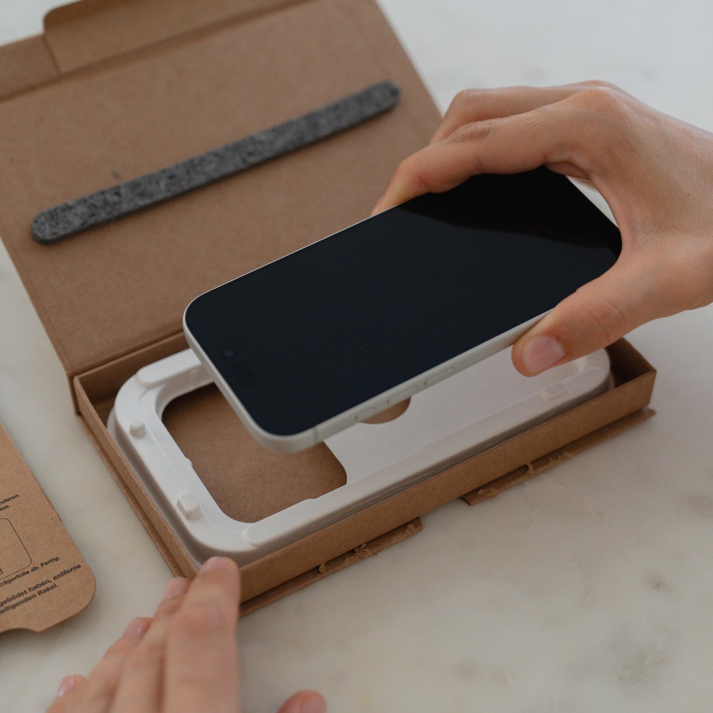 Hände beim Unboxing eines Smartphones mit einer EZ.INSTALL Box und arktis 2er Set iPhone 17 Pro Max REVOLUTION Glas-Displayschutz aus recycelter Verpackung auf einer weißen Fläche.