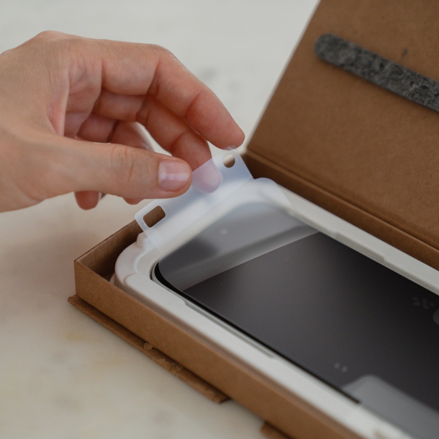 Eine Hand hält eine Kunststoffabdeckung, bereit zum Anbringen des arktis 2er Set iPhone 17 Pro Max REVOLUTION Glas-Displayschutzes mit der EZ.INSTALL Box für einfachen Bildschirmschutz.