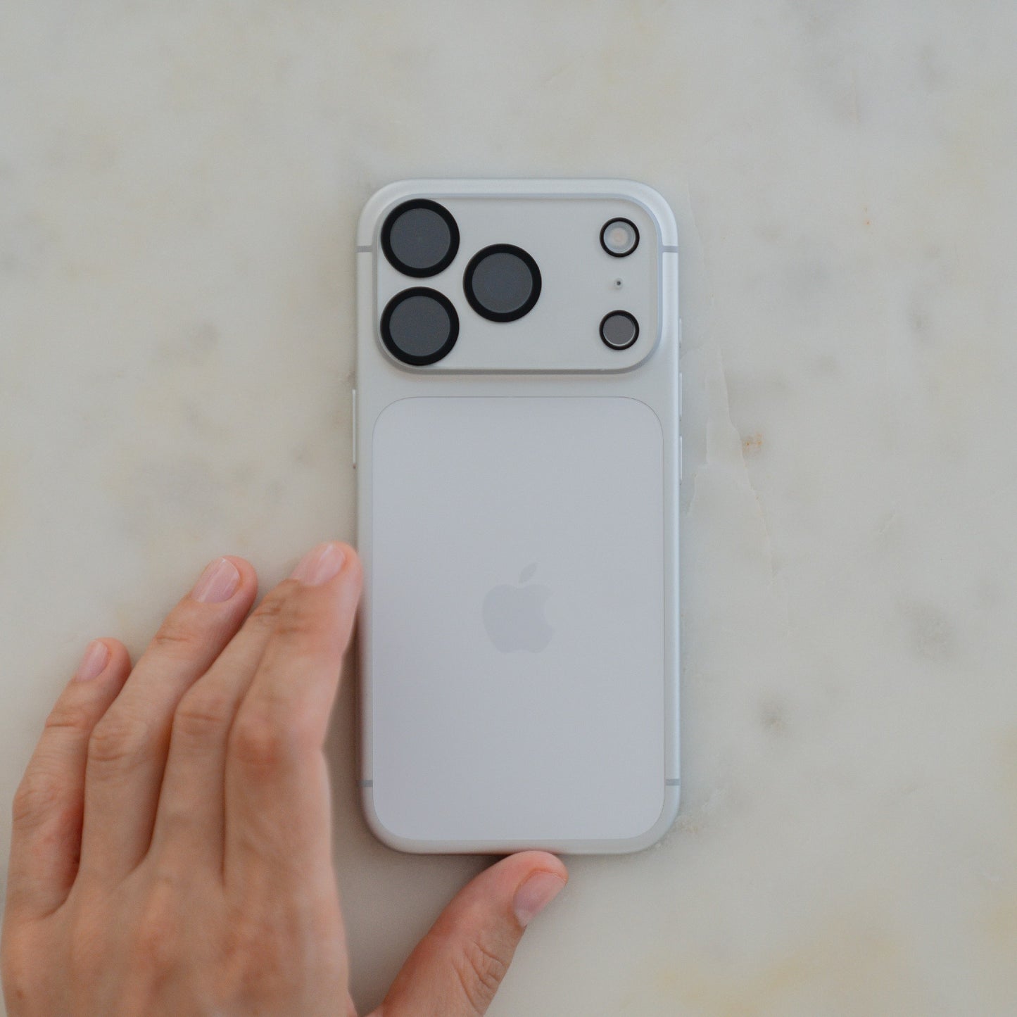 Eine Hand berührt ein weißes iPhone mit drei Kameras, geschützt durch den arktis iPhone 17 Pro Max Kamera Protector von arktis, auf einer hellen Marmorfläche.