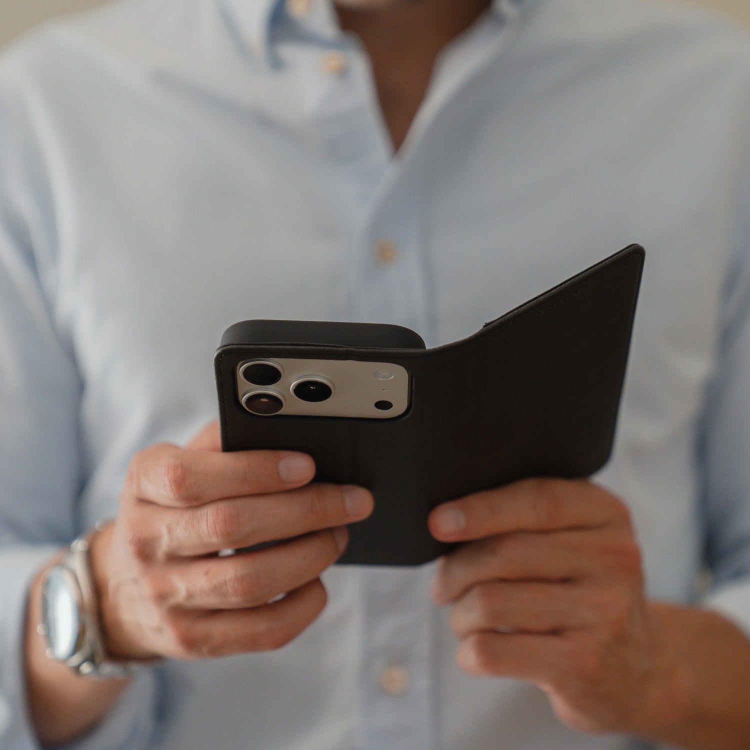 Eine Person in einem blauen Hemd hält ein iPhone 17 Pro in der schwarzen arktis iPhone 17 Pro Book Tasche, die sowohl Stil als auch Schutz hervorhebt.