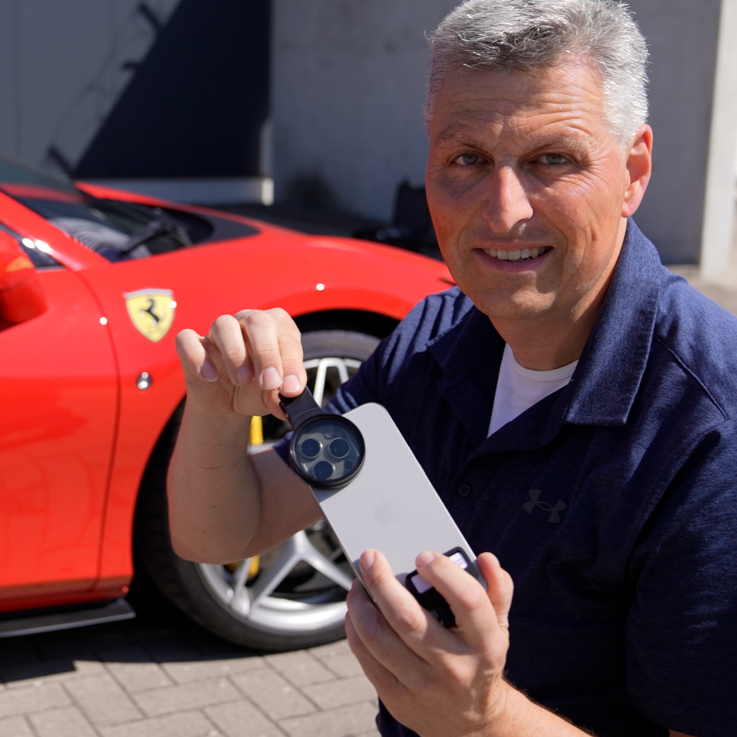 Ein Mann hält an einem sonnigen Tag sein mit dem Wolffilms CarLens PRO Polfilter ausgestattetes Smartphone neben einen roten Ferrari und hält den Moment fest – perfekt für Autovideo-Enthusiasten.
