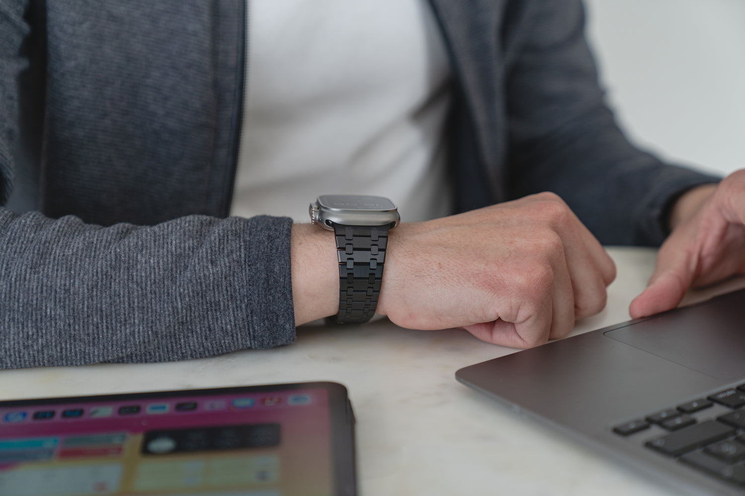 Eine Person, die ein elegantes Arktisband Edelstahlarmband „Symphony“ für die Apple Watch mit Butterfly-Schließe trägt, arbeitet an einem Laptop, während in der Nähe ein Tablet auf dem Tisch liegt.