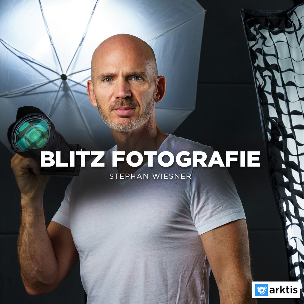 Ein glatzköpfiger Mann in weißem T-Shirt steht mit einem Kamerablitz in einem Studio – ideal für Blitz- oder Portraitfotografie. Hinter ihm befinden sich ein Schirmreflektor und eine Softbox. Der Text zeigt Fotografieren mit Blitz Stephan Wiesner; Arktis-Logo unten rechts.