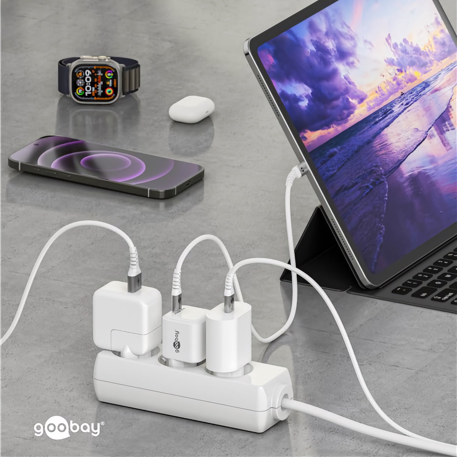 Ein Tablet, ein Smartphone und eine Smartwatch in der Nähe einer Steckdosenleiste mit einem Goobay USB-C PD GaN Schnellladegerät Nano 30 W zum Aufladen mehrerer Geräte auf einer grauen Fläche.