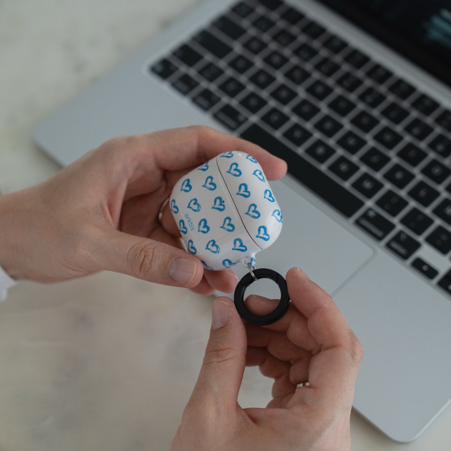 Hände halten eine Arktis AirPods Pro 3 Hülle – Blue Hearts-Hülle neben der Tastatur eines Laptops auf einer weißen Fläche.