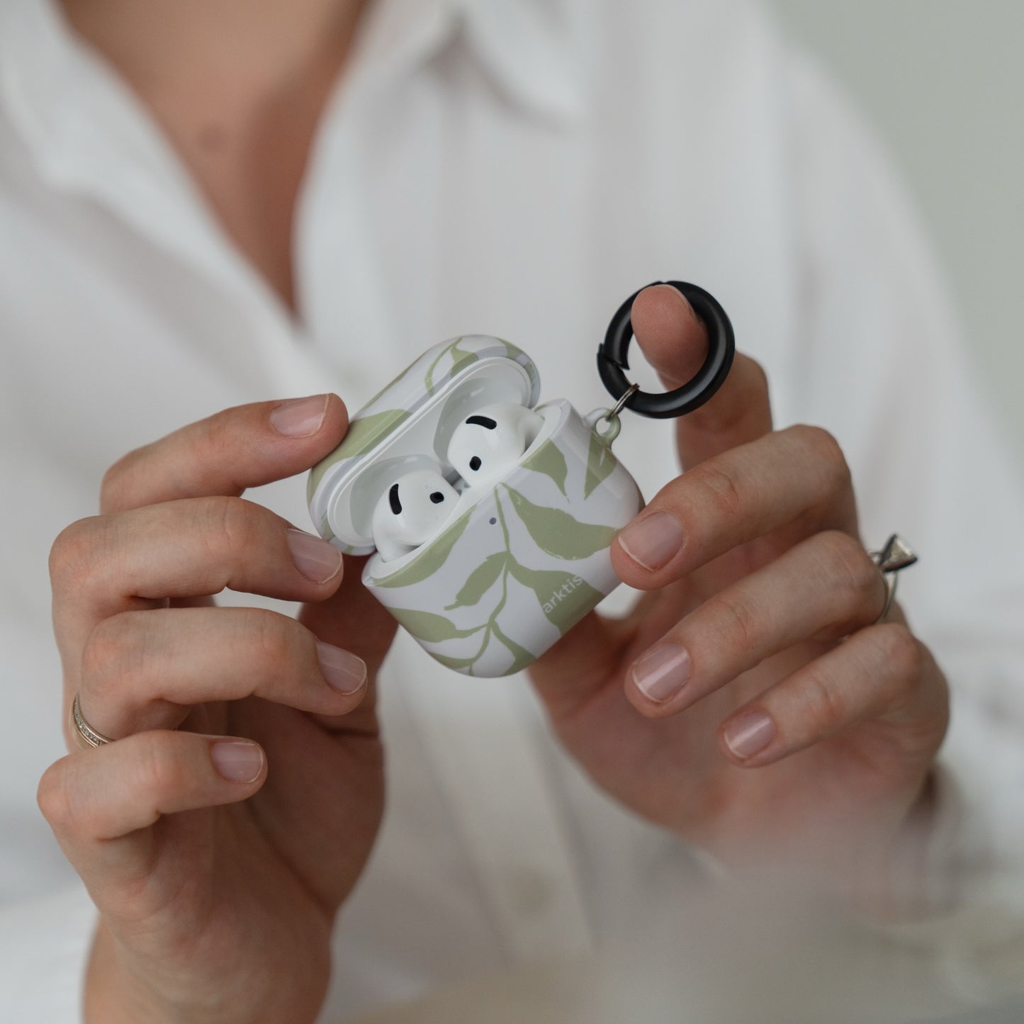 Hände, die eine geöffnete, weiße Arktis AirPods 5-Hülle – Botanic – mit grünem Blattmuster und einem schwarzen Karabinerring halten.