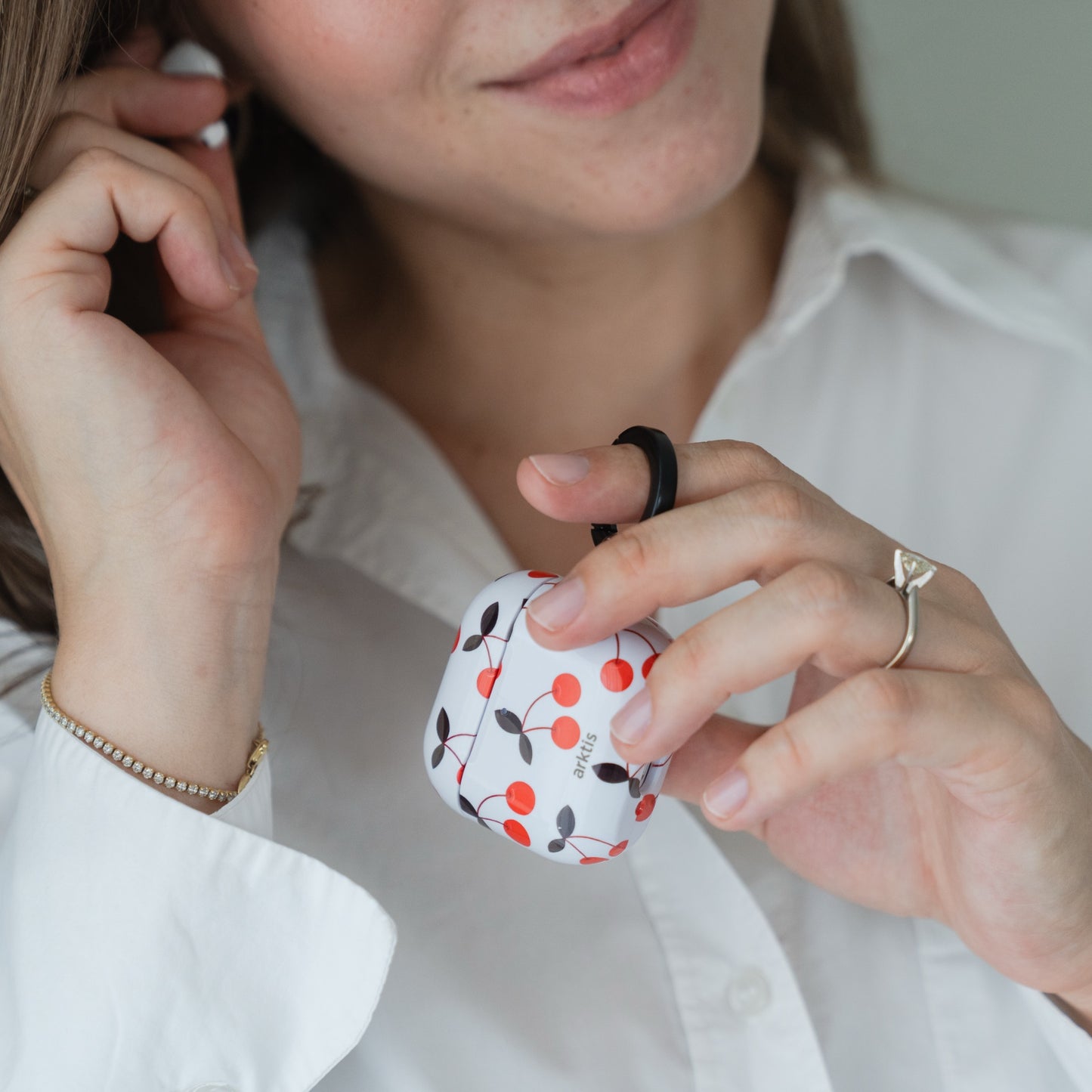 Eine Frau in einem weißen Hemd lächelt, während sie die Arktis AirPods 5 Hülle – Cherry in der Hand hält und sich einen Ohrhörer ins Ohr steckt.