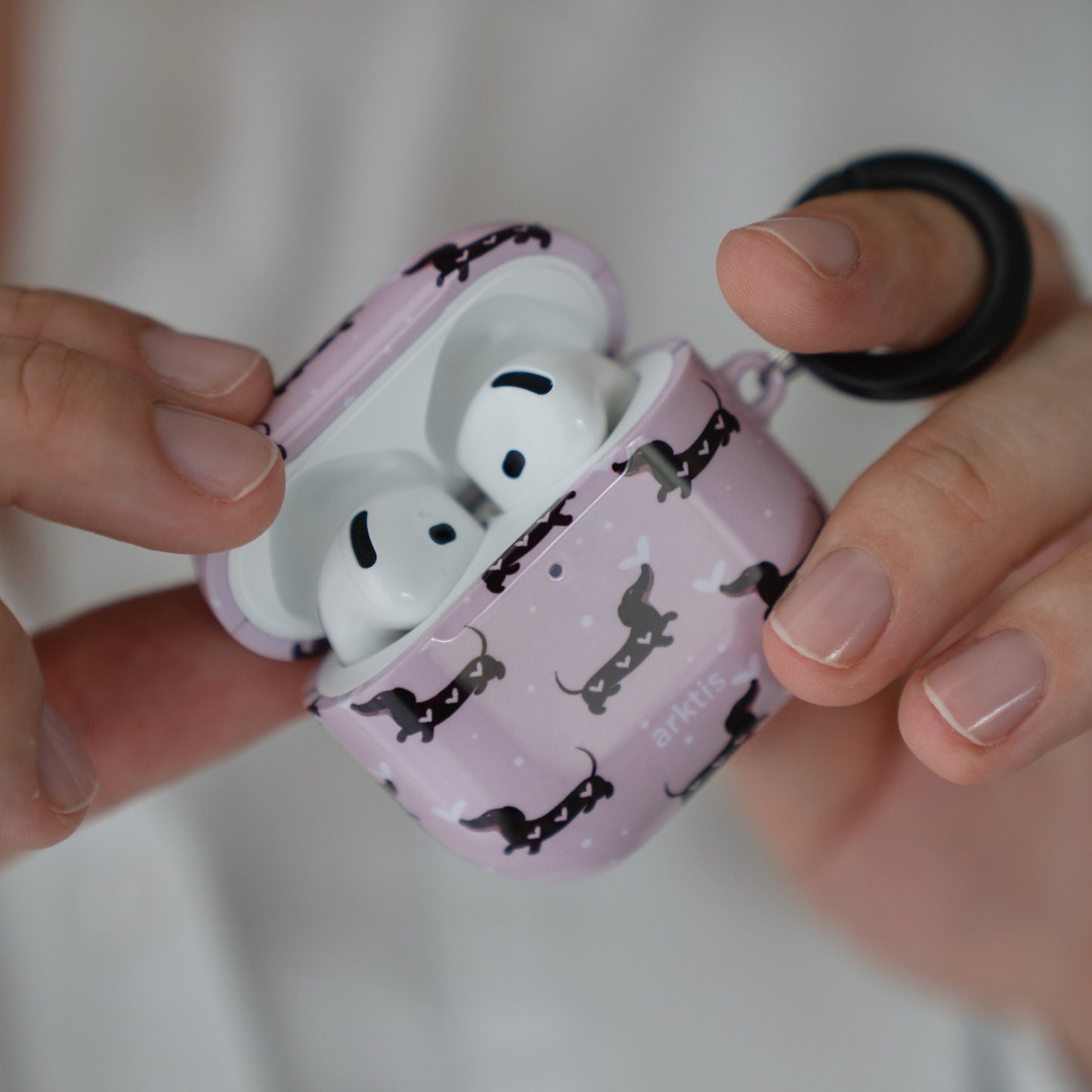 Hände, die eine geöffnete Arktis AirPods 5-Hülle halten – Dackel Love-Hülle mit einem rosa Dackelmuster.
