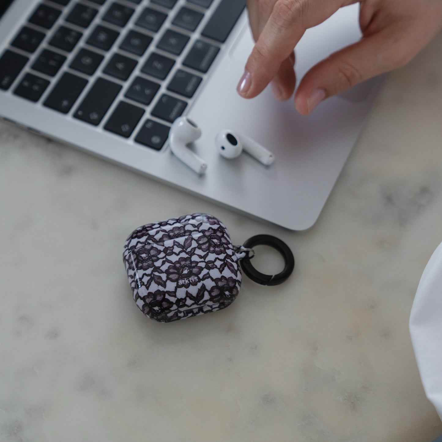 Eine Hand greift nach den AirPods neben einem Laptop, auf einer Marmoroberfläche liegt eine Arktis AirPods 5 Hülle – Dark Rose.