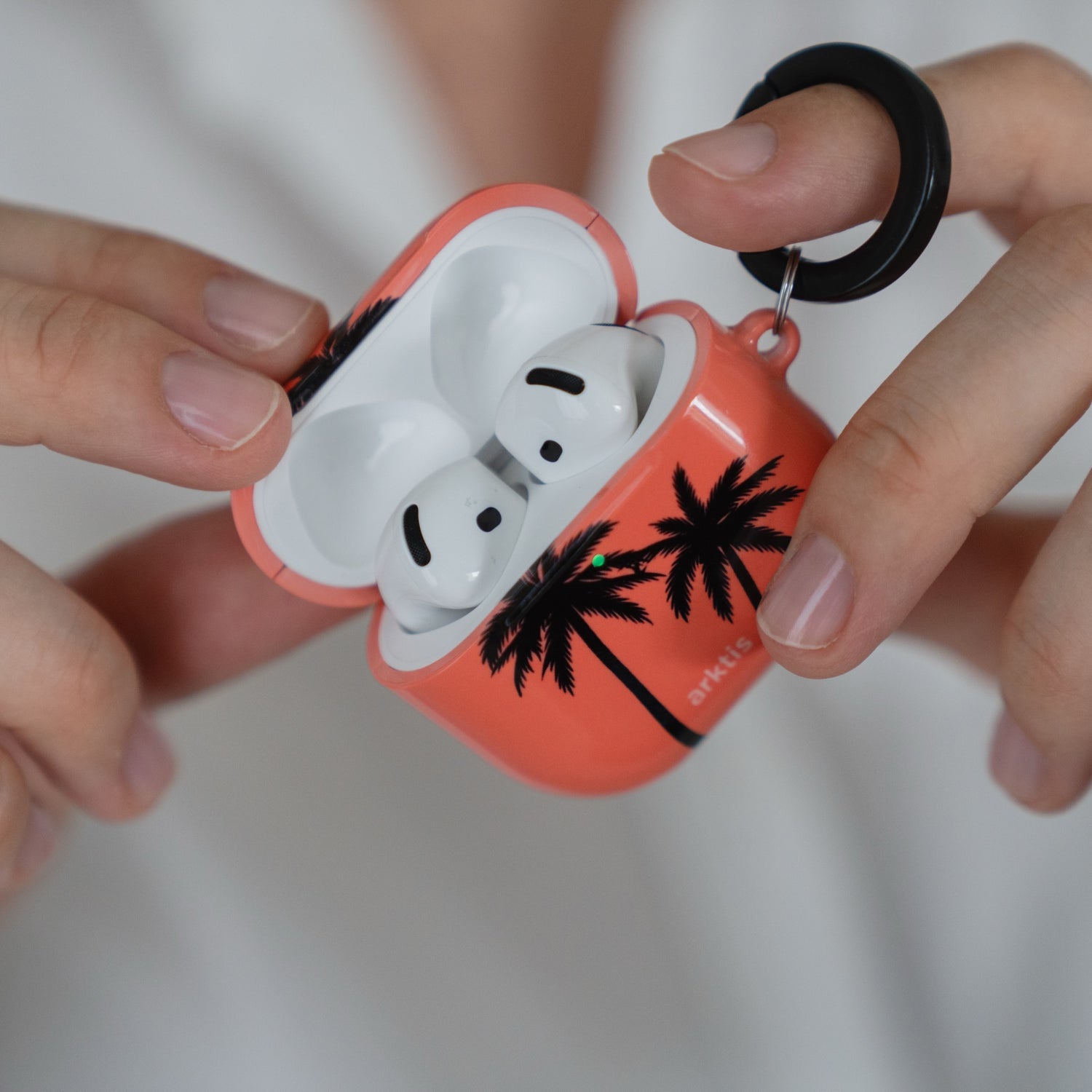 Hände, die eine geöffnete Arktis AirPods Pro 3-Hülle in der Farbe Coral Coast mit schwarzem Palmenmuster und einem kleinen Karabinerhalter halten.