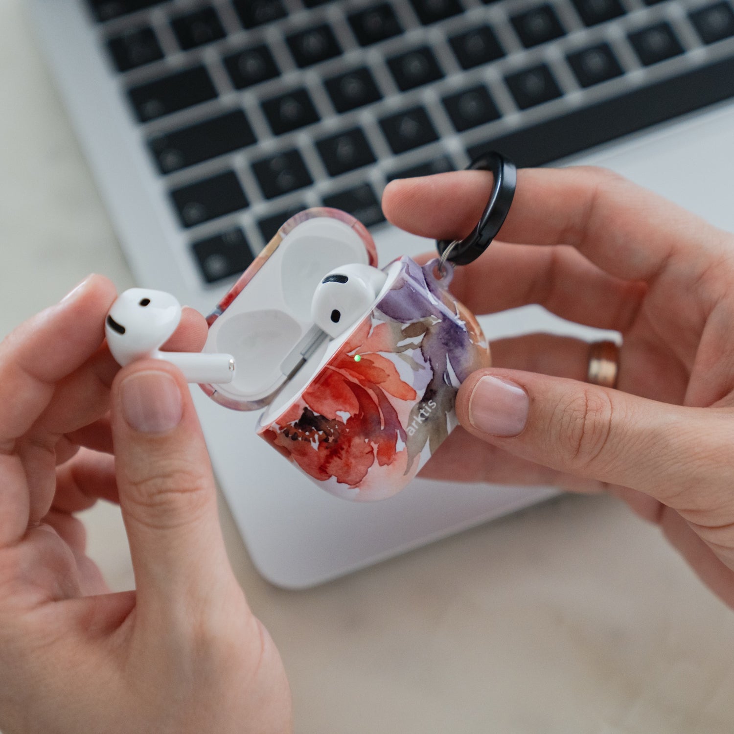 Hände, die eine geöffnete Arktis AirPods 5-Hülle halten – Ladecase mit Blumenmuster, aus dem ein Ohrhörer entnommen wurde.