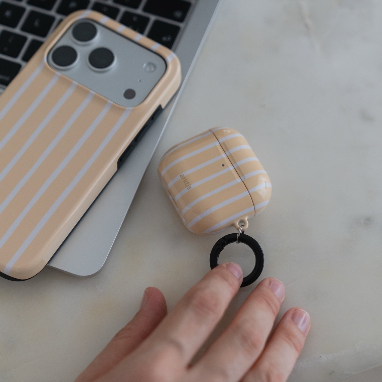 Eine Hand berührt den schwarzen Ringclip einer Arktis AirPods 5-Hülle – Sunny Stripes – und die dazu passende Handyhülle mit gelben Streifen.