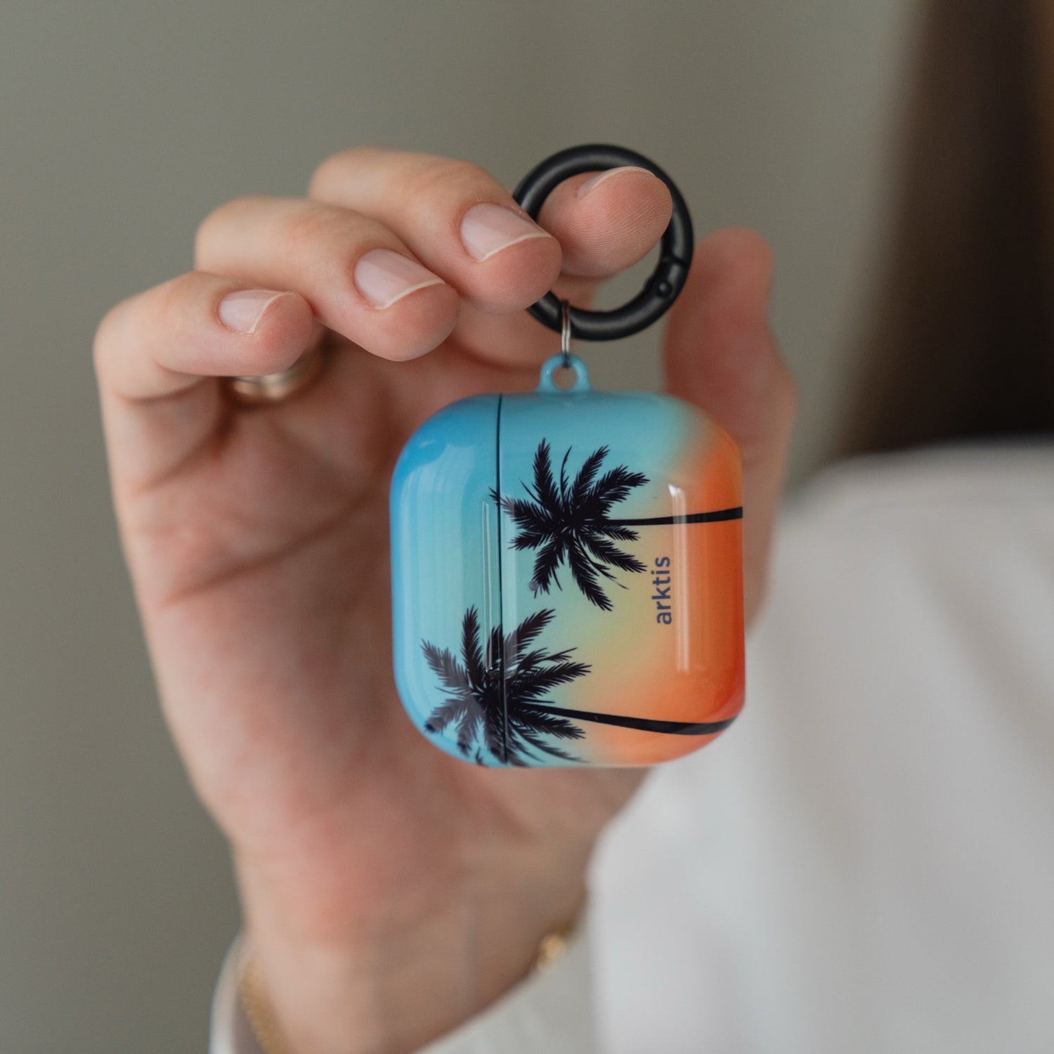 Eine Hand, die eine Arktis AirPods 5-Hülle hält – Sunset Beach mit einem Aufdruck von Sonnenuntergang und Palmen.