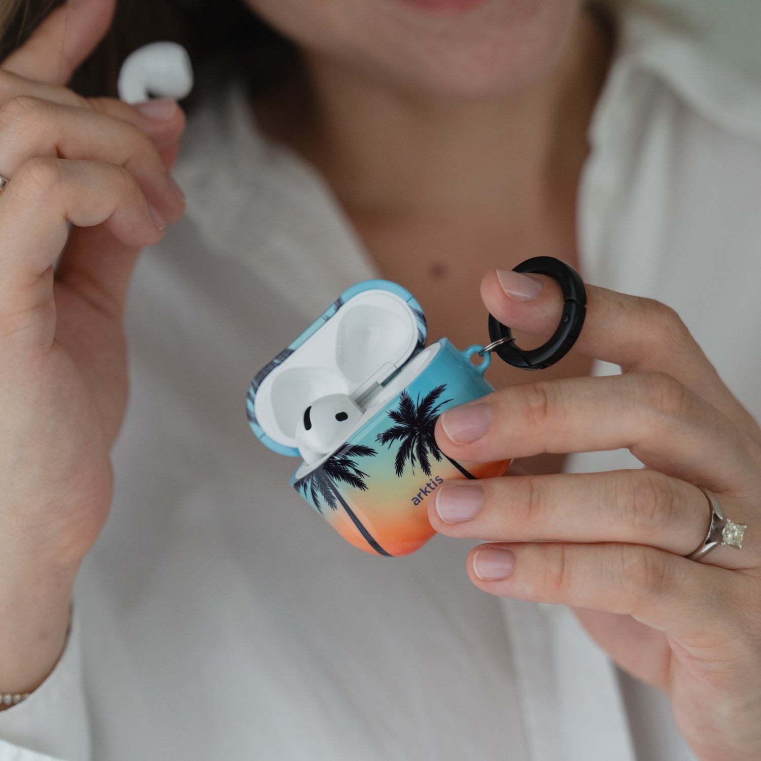 Open arktis AirPods 5 Hülle – Sunset Beach-Hülle mit einem Aufdruck, der einen Sonnenuntergang mit Palmen zeigt, und einem schwarzen Befestigungsring.