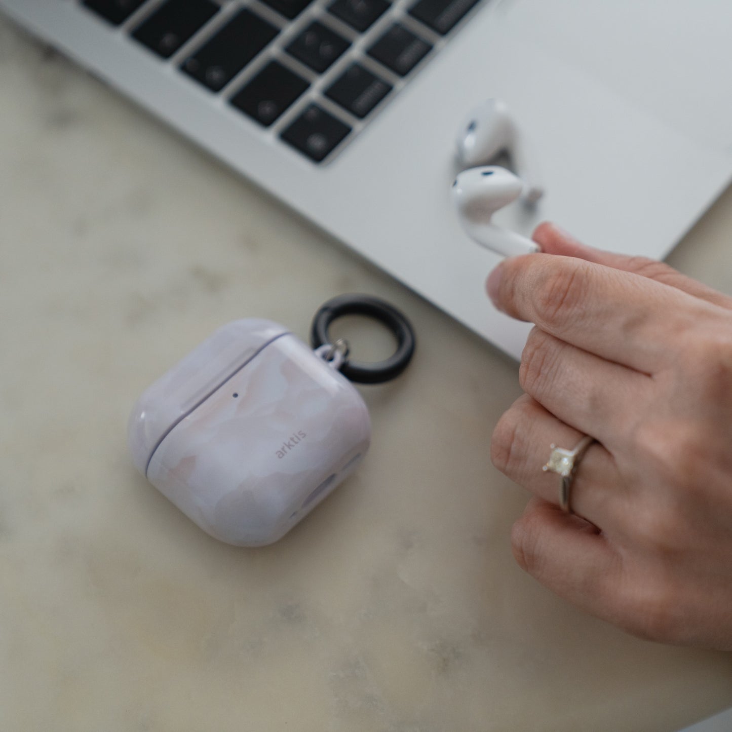 Eine Hand, die kabellose Ohrhörer neben einer Arktis AirPods 5-Hülle hält – einem Marble Flower-Ladeetui mit schwarzem Ring.