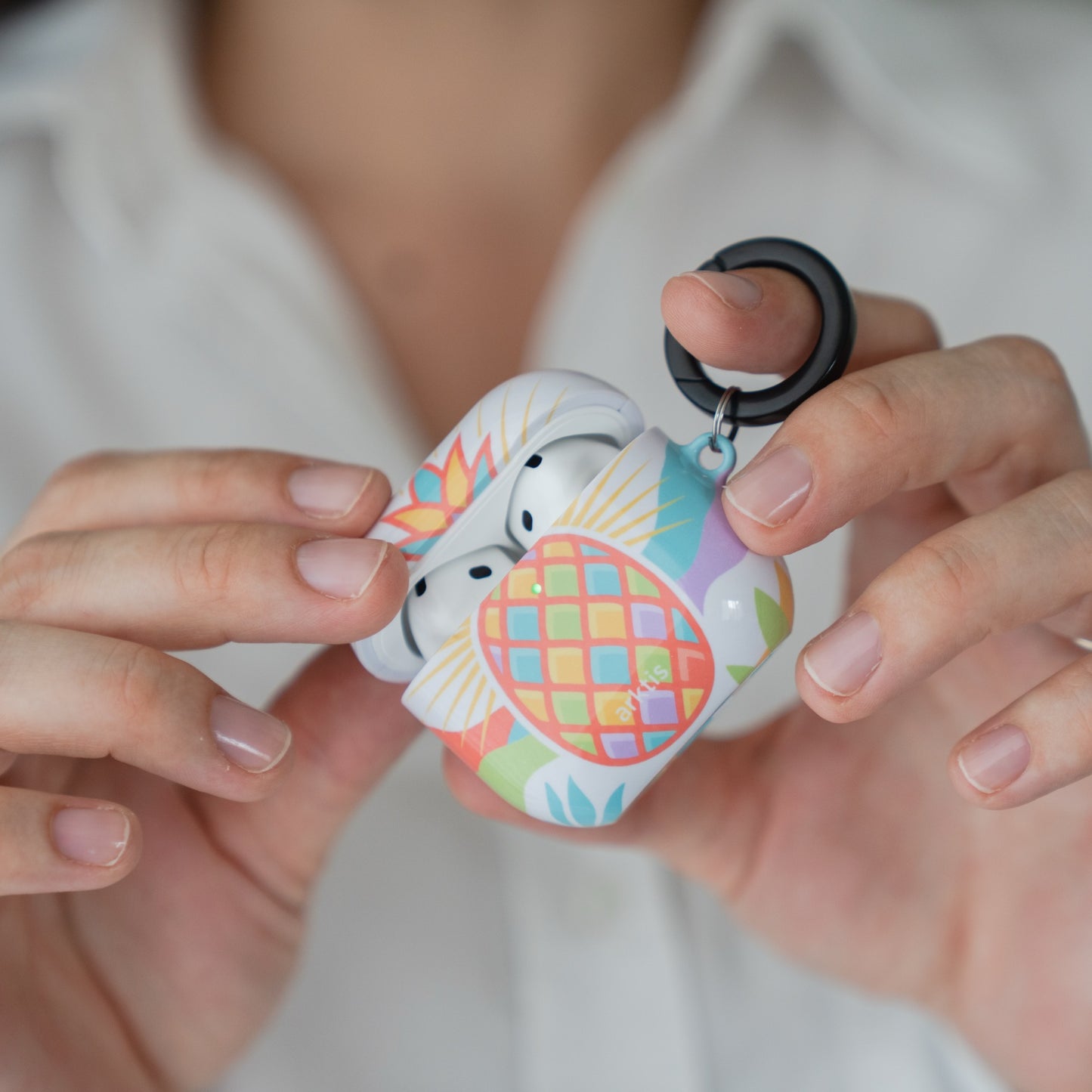 Person, die eine geöffnete Arktis AirPods 5 Hülle – Tropic Pineapple-Schutzhülle mit einem daran befestigten schwarzen Karabiner in der Hand hält.