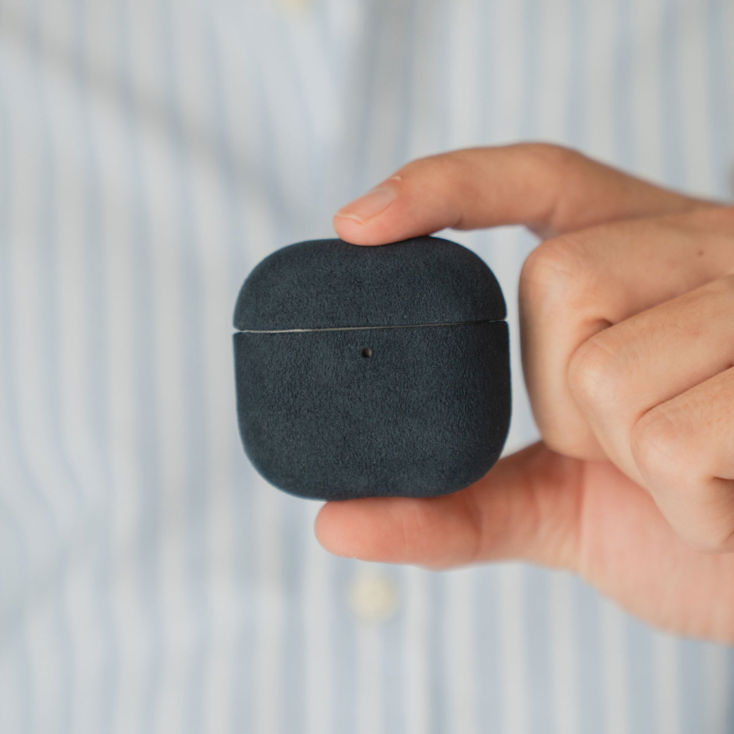Eine Hand hält eine Arktis AirPods Pro 3-Hülle aus Alcantara vor einer Person in einem blau gestreiften Hemd.