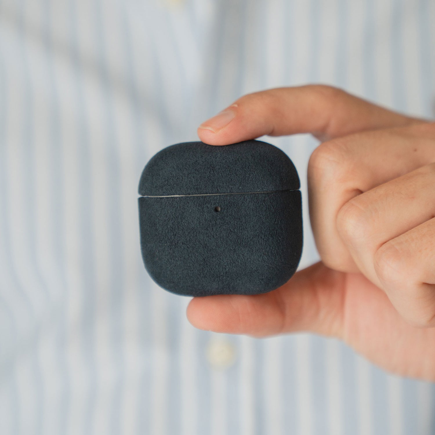 Eine Hand hält eine Arktis AirPods Pro 3-Hülle aus Alcantara vor einer Person in einem blau gestreiften Hemd.