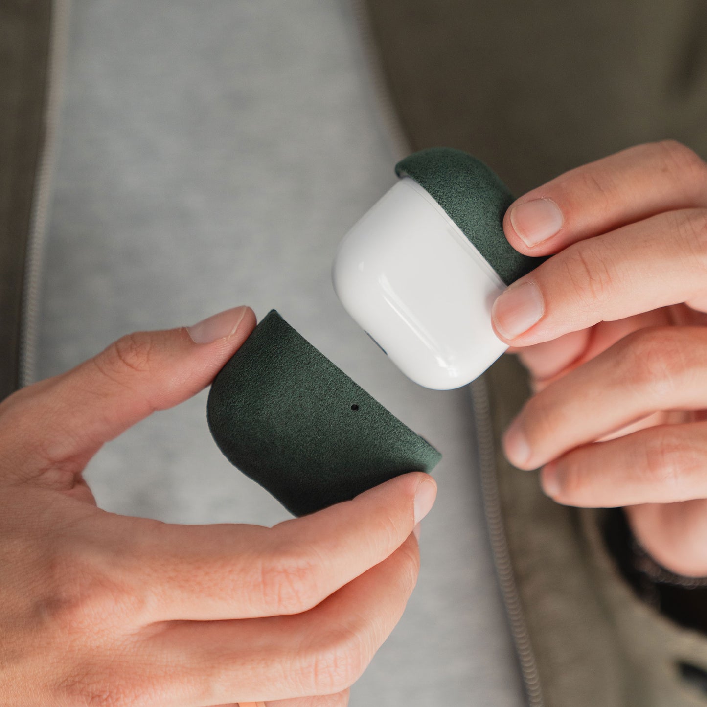 Hände halten eine geöffnete Arktis AirPods Pro 3-Hülle aus Alcantara in Dunkelgrün und Weiß vor einem neutralen Hintergrund.