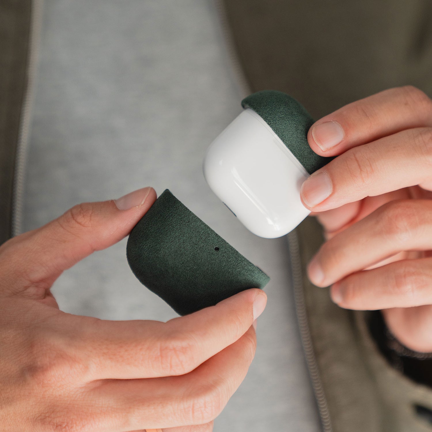 Hände halten eine geöffnete Arktis AirPods Pro 3-Hülle aus Alcantara in Dunkelgrün und Weiß vor einem neutralen Hintergrund.