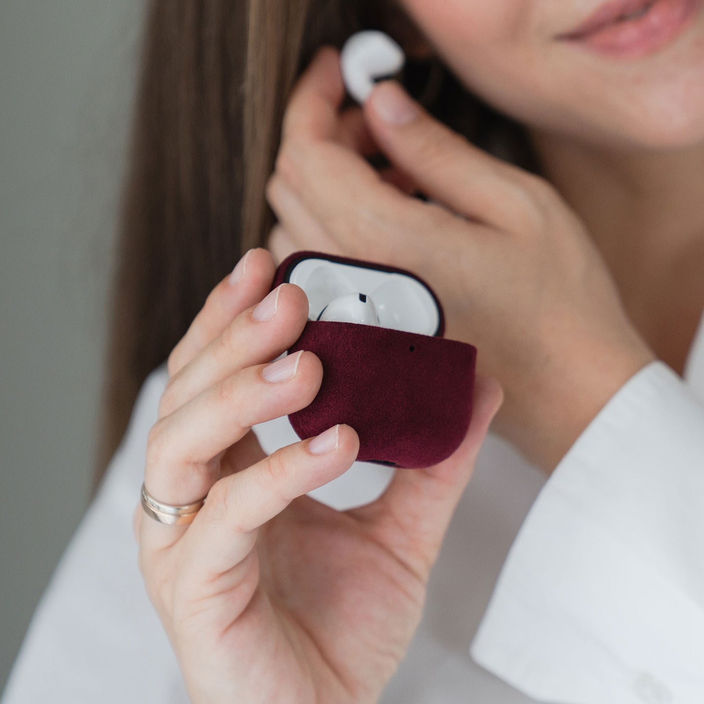 Eine Person steckt sich Ohrhörer in die Ohren, hält dabei eine burgunderrote Arktis AirPods Pro 3-Hülle aus Alcantara in der Hand und trägt einen silbernen Ring.