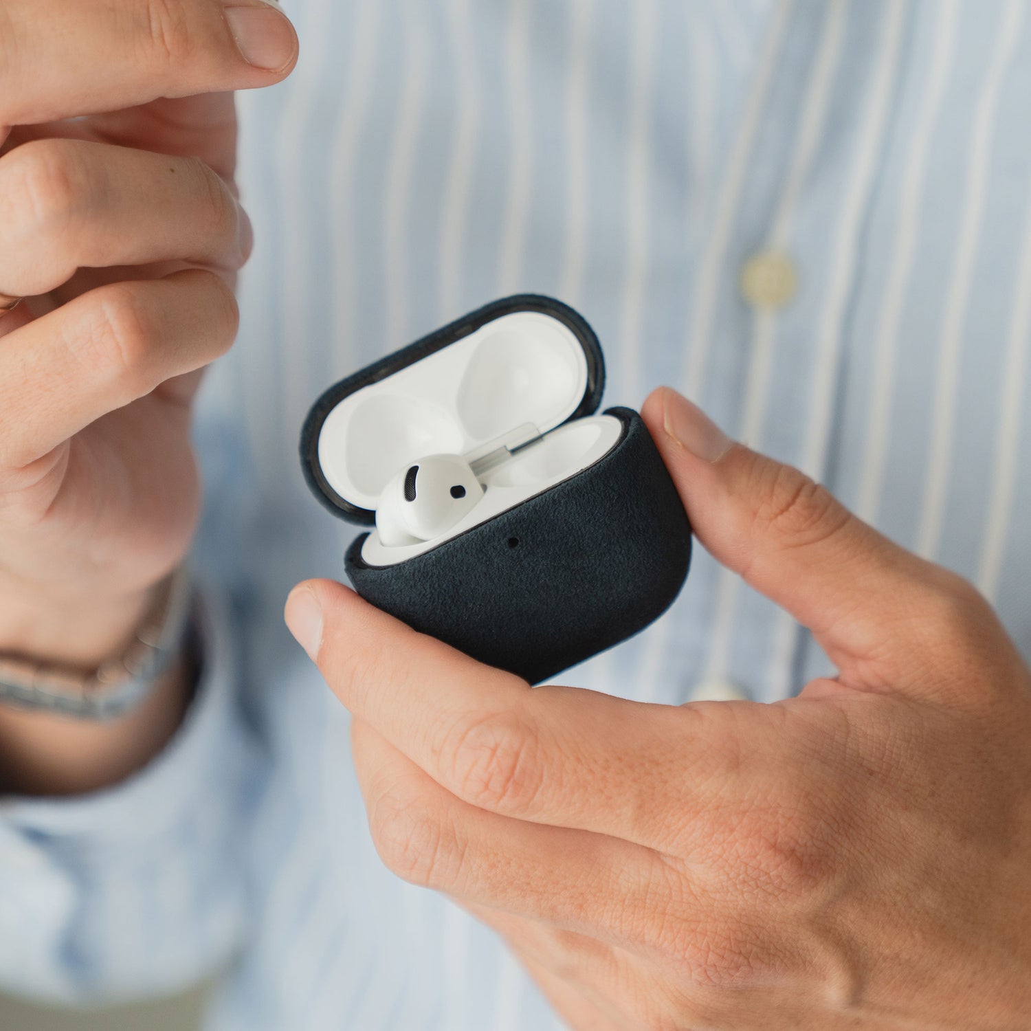 Jemand hält eine Arktis AirPods Pro 3-Hülle aus Alcantara mit einem Ohrhörer in der Hand und trägt ein blau gestreiftes Hemd.