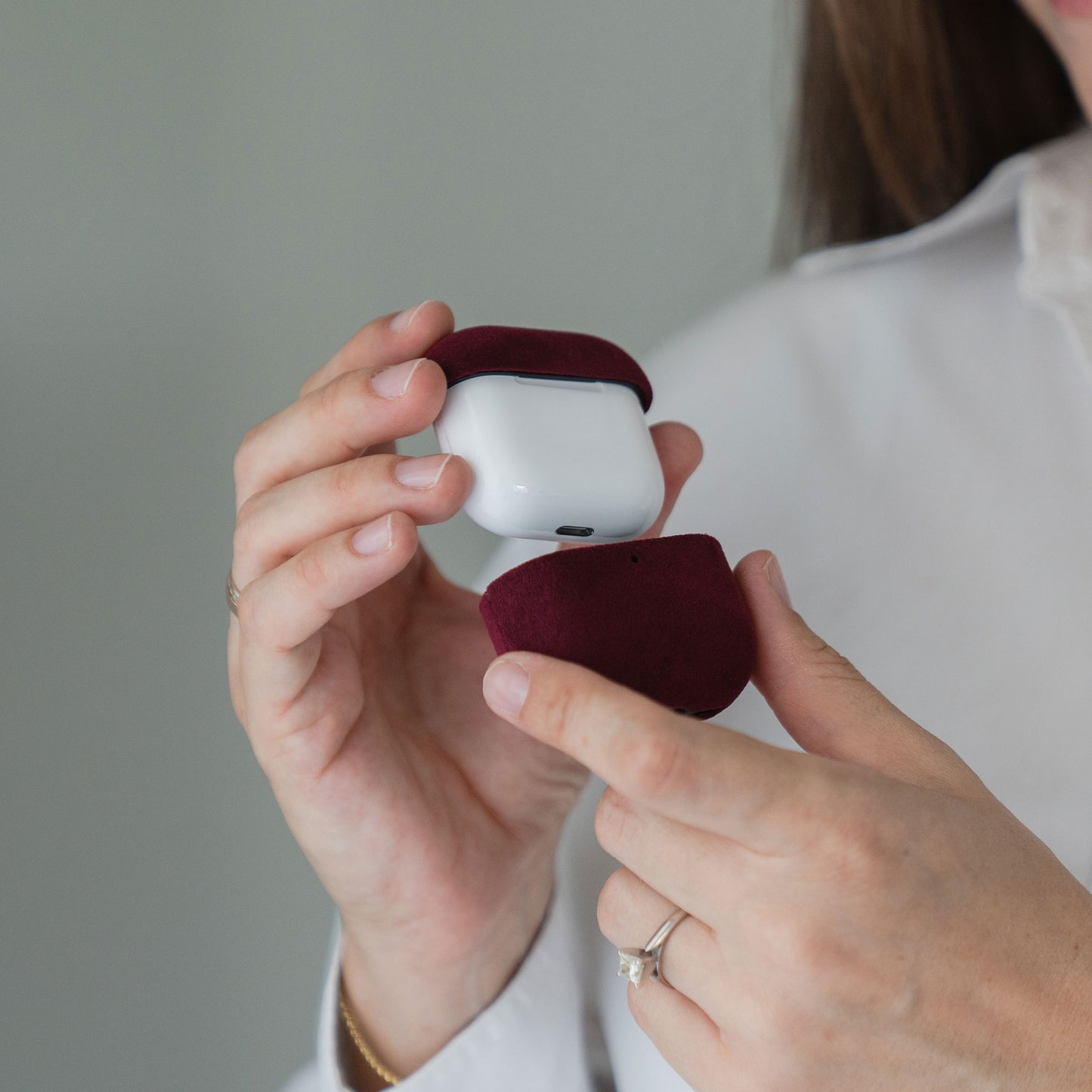 Eine Person hält eine burgunderrote Arktis AirPods Pro 3-Hülle aus Alcantara mit darin befindlichen AirPods in der Hand und trägt ein weißes Hemd sowie einen Ring.