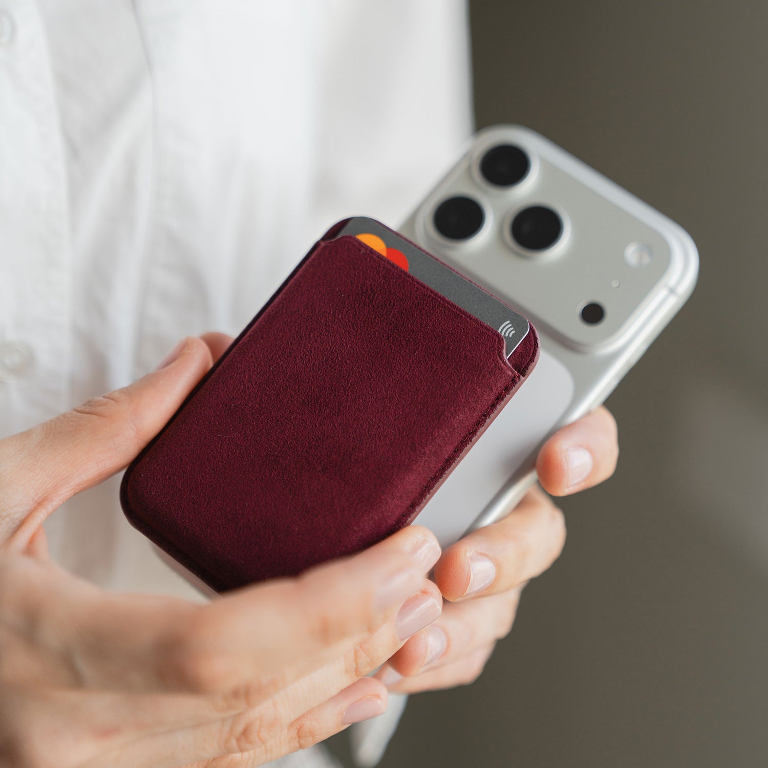 Eine Person bringt einen arktis iPhone MagSafe Kartenhalter aus Alcantara in Burgunderrot auf der Rückseite eines silbernen iPhones an.