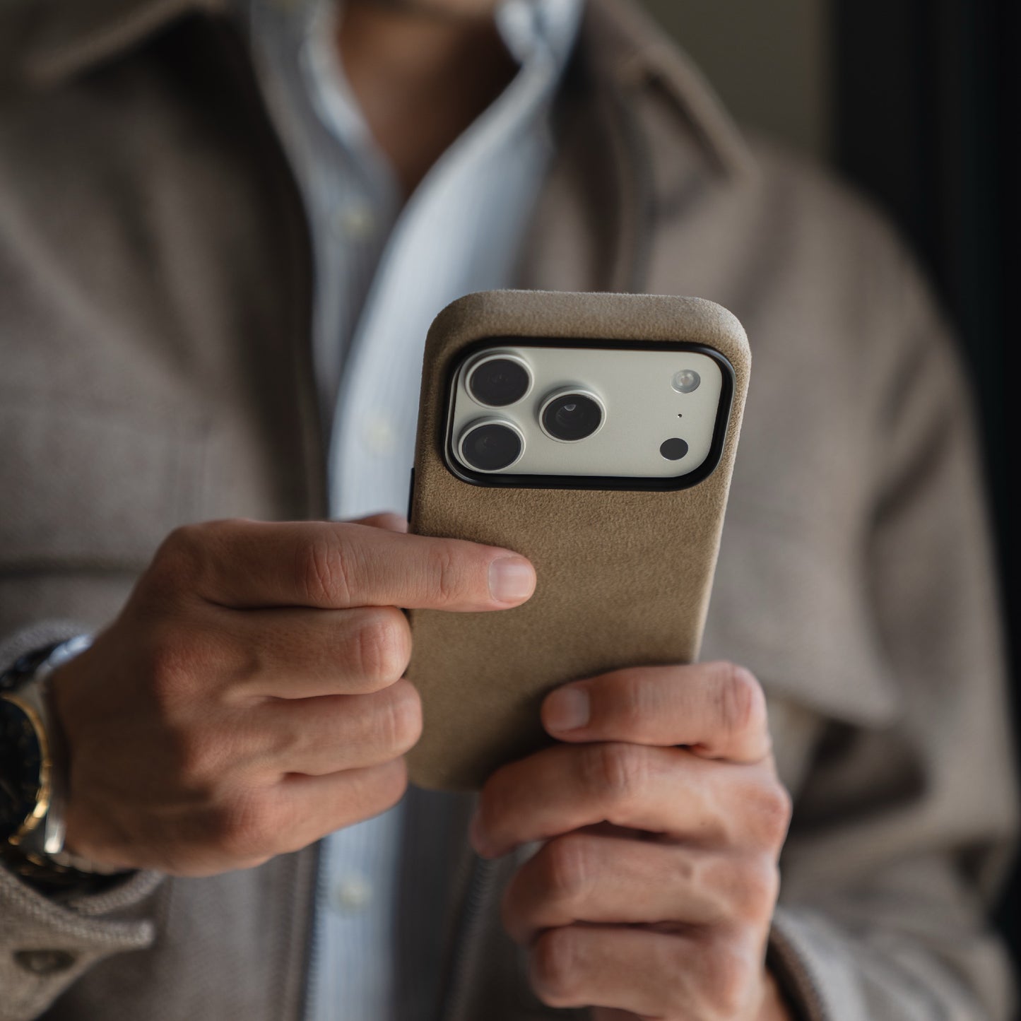 Eine Person hält ein Smartphone mit einer arktis iPhone 17 Pro Hülle aus Alcantara, made in Italy, und trägt eine braune Jacke und ein hellblaues Hemd.