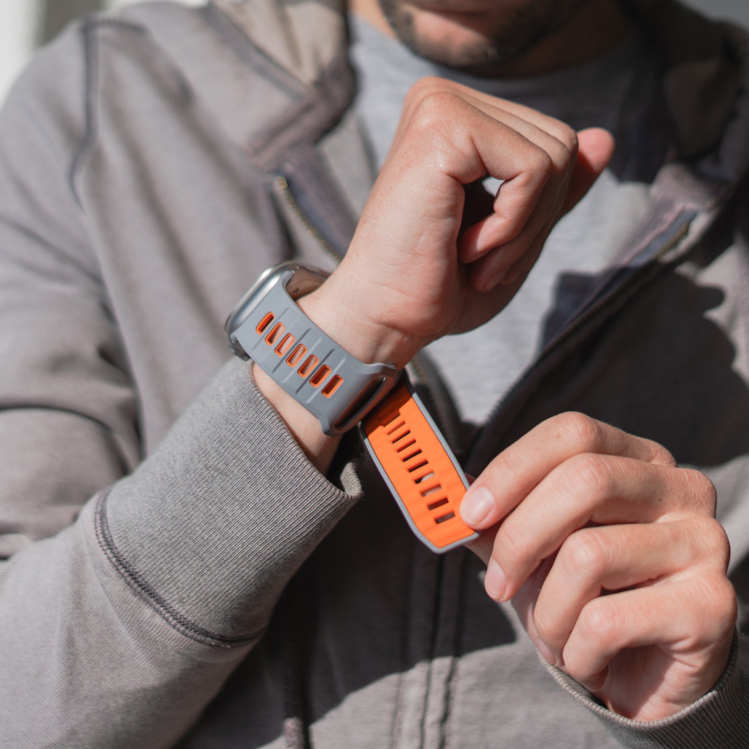 Eine Person in einem grauen Hoodie befestigt das arktisband Apple Watch Silikonarmband „Runner“ (orange/grau) an ihrem Handgelenk. Das Bild hebt die atmungsaktive Bandstruktur und das stylische Design hervor, wobei nur die Zeiger und die Uhr sichtbar sind.
