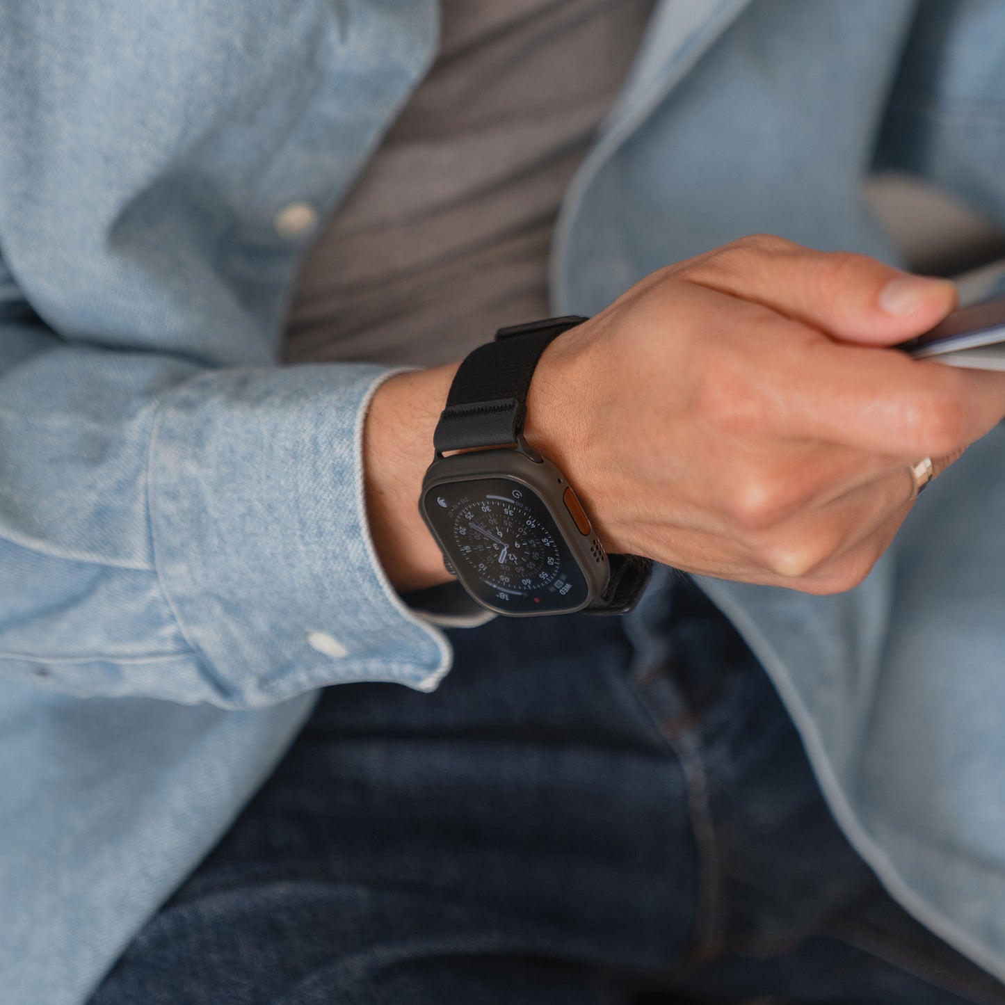 Eine Person trägt das arktisband Apple Watch Armband "Terra" von arktisband - ein schwarzes, extrabreites Nylonarmband - gepaart mit einem blauen Jeanshemd, während sie ein Smartphone in ihrer rechten Hand hält.