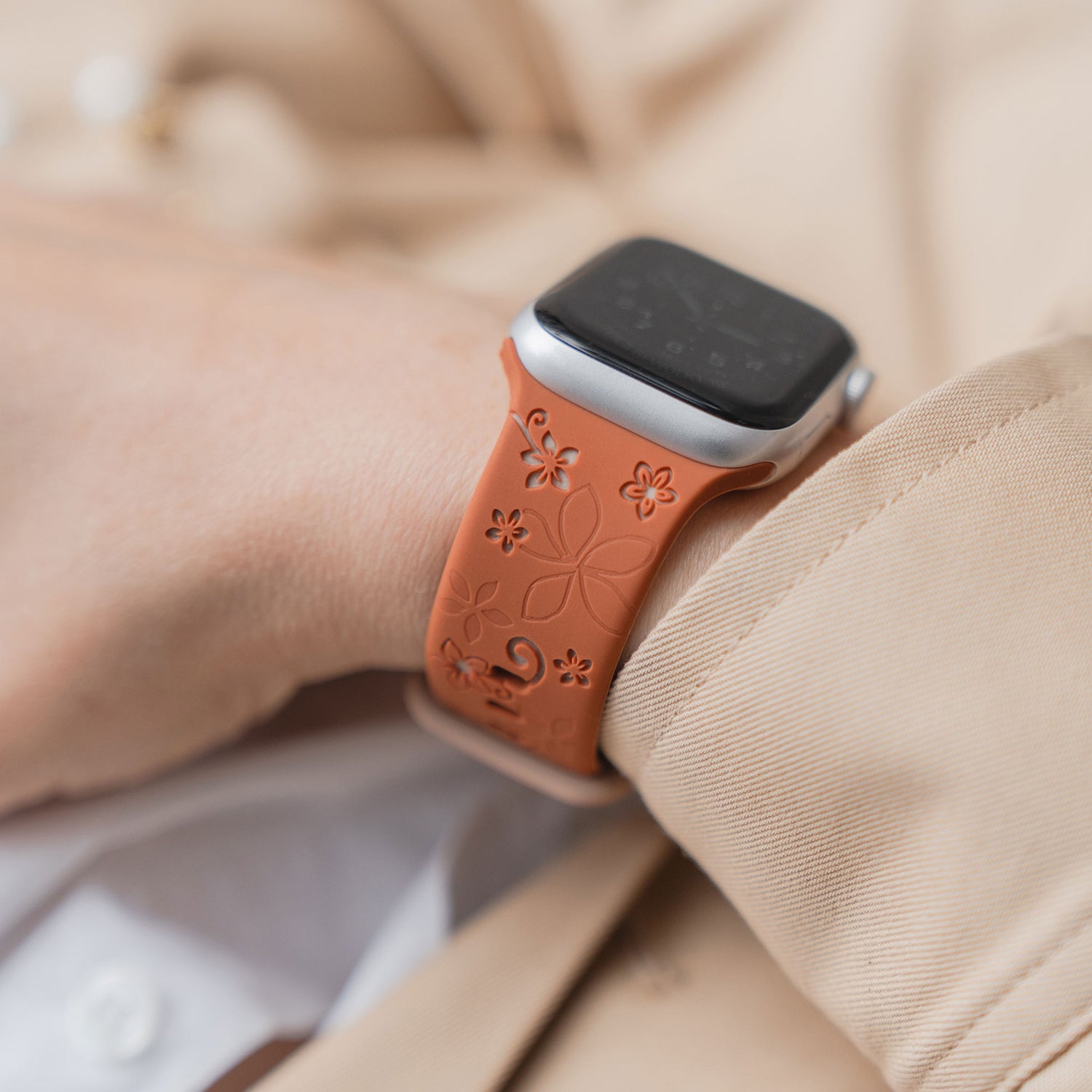 Nahaufnahme eines Handgelenks, das das arktisband Apple Watch Silikonarmband "Bloom" mit einem orangefarbenen Blumenmuster trägt, gepaart mit einer beigen Hülle.
