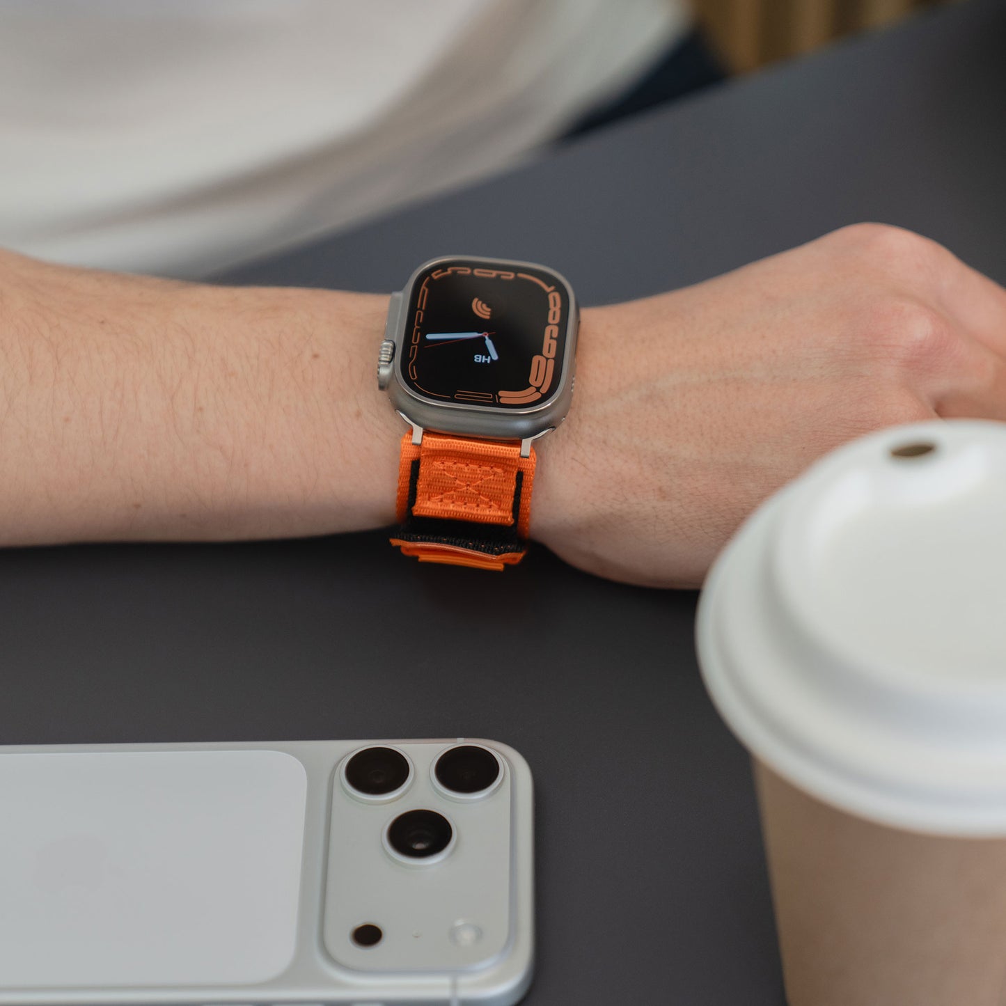 Eine Person, die ein orangefarbenes arktisband Apple Watch Outdoor Armband "Forge" von arktisband trägt, sitzt an einem Tisch mit einem weißen Smartphone und einer Kaffeetasse.
