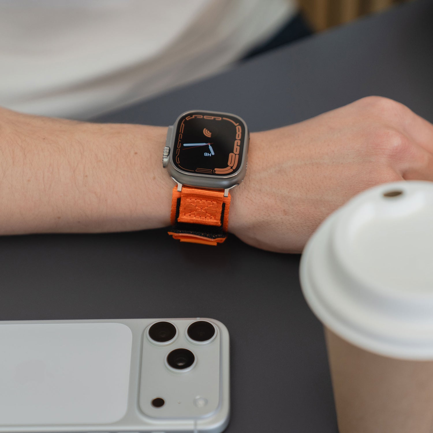 Eine Person, die ein orangefarbenes arktisband Apple Watch Outdoor Armband "Forge" von arktisband trägt, sitzt an einem Tisch mit einem weißen Smartphone und einer Kaffeetasse.