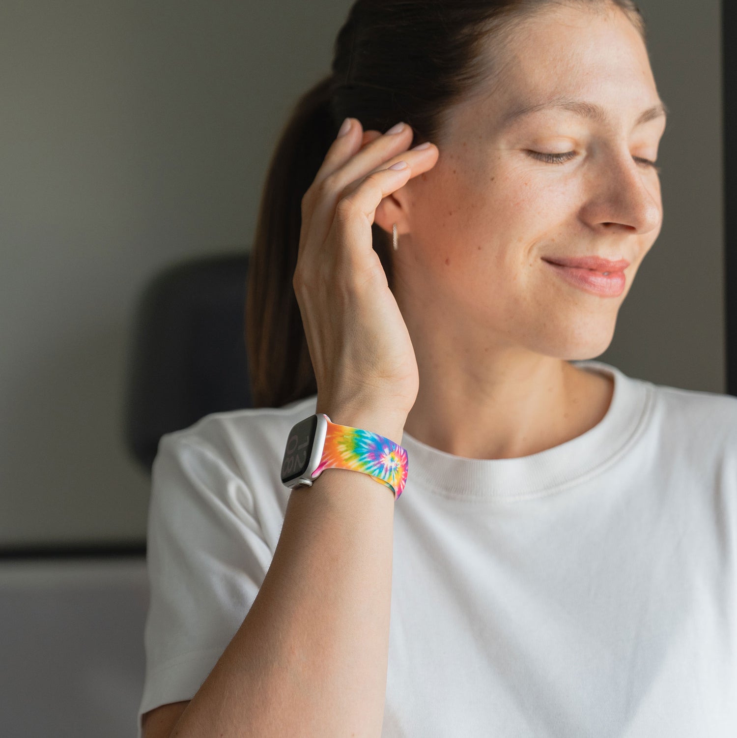 Lächelnde Frau im weißen Hemd trägt das arktisband Batik Rainbow Apple Watch Armband von arktisband mit buntem Tie-Dye Design und berührt dabei ihr Ohr.