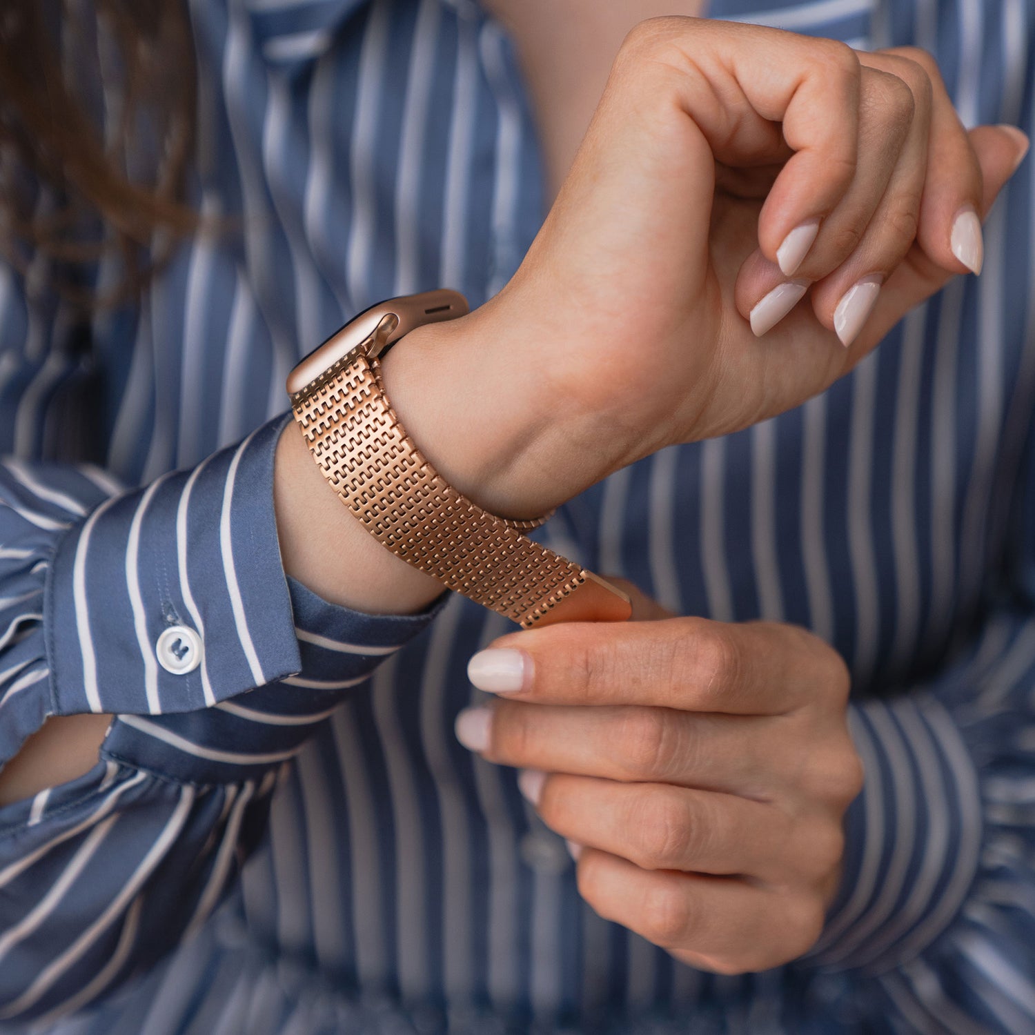 Eine Frau im gestreiften Shirt befestigt das roségoldene arktisband Apple Watch Edelstahl Armband „Aurora“ mit Magnetverschluss an ihrem Handgelenk und verpasst ihrer Apple Watch so ein stylisches und sicheres Upgrade von arktisband.