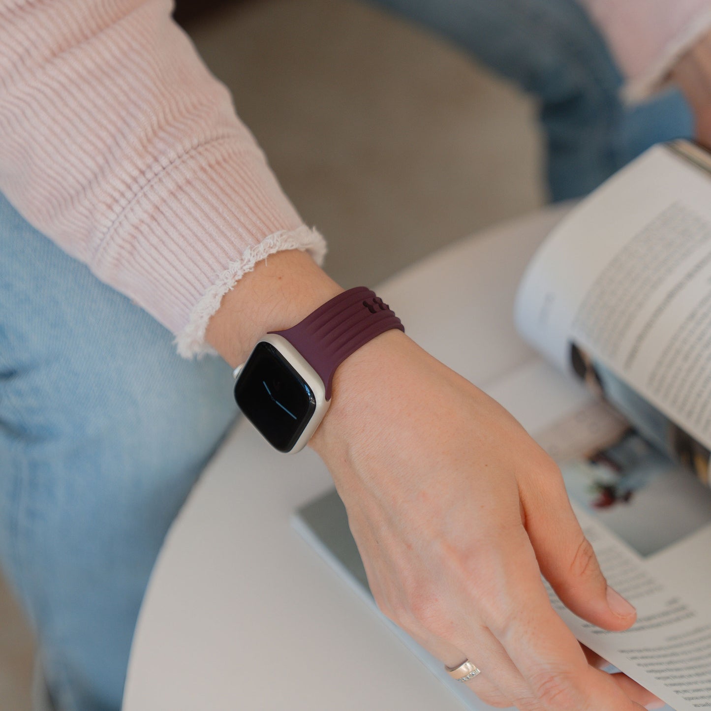 Eine Person in einem rosafarbenen Pullover und einem arktisband Apple Watch Armband "Skandi" von arktisband liest eine Zeitschrift an einem weißen Tisch, der den Skandi-Designstil verkörpert.