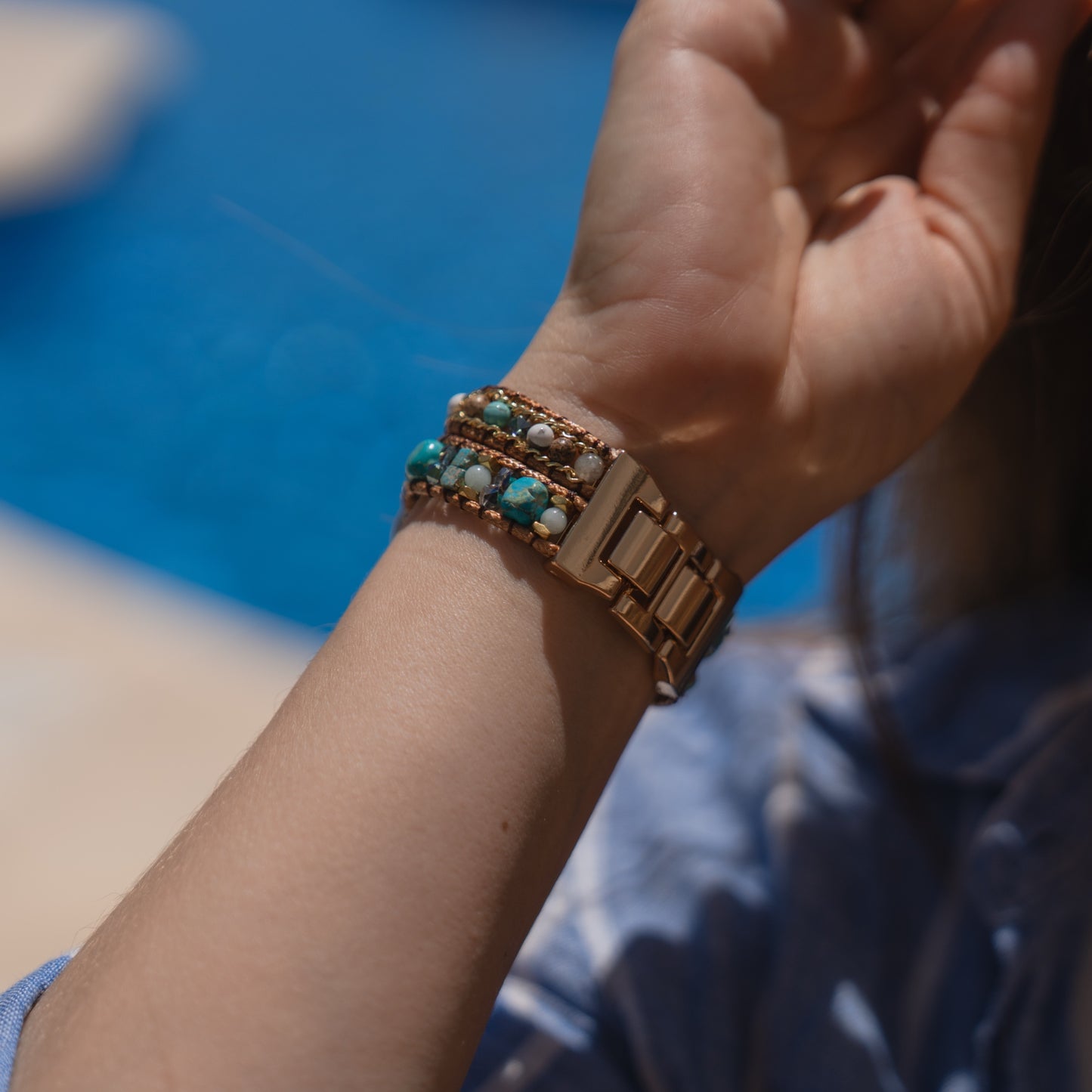 Ein Handgelenk mit einer goldenen Uhr und Armbändern aus türkisfarbenen Perlen, an dem das Apple Watch-Armband „Sunset Spirit“ von arktisband zu sehen ist, neben einem blauen Swimmingpool.