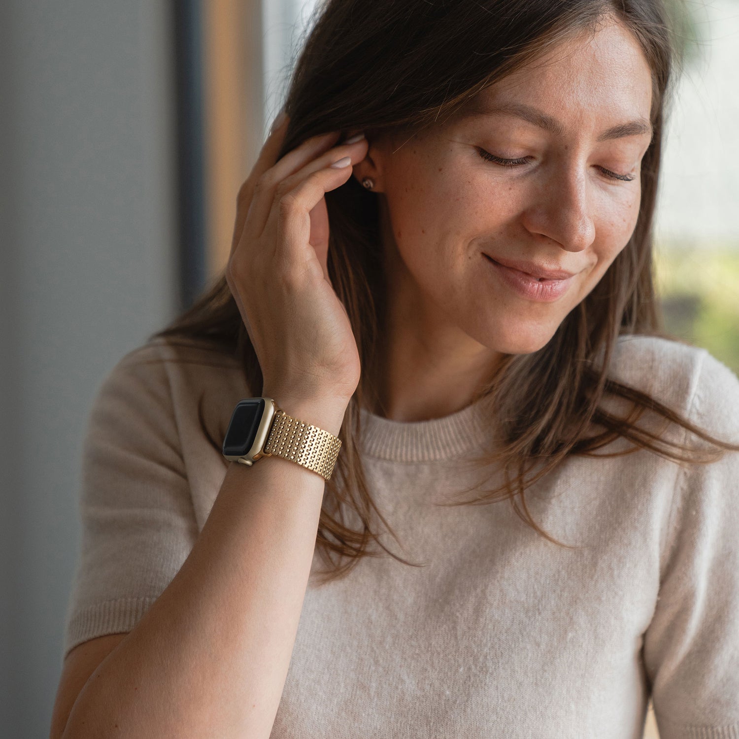 Eine Frau lächelt mit geschlossenen Augen, berührt ihr Ohr und trägt ein beiges Oberteil sowie das goldene Arktisband Apple Watch Edelstahlarmband „Aurora“ mit elegantem Magnetverschluss.