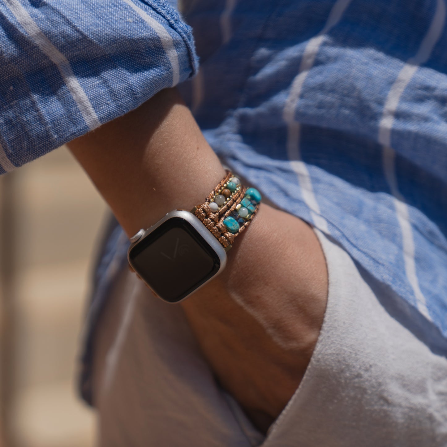 Eine Person, die ein blau gestreiftes Hemd und ein Apple Watch-Armband „Sunset Spirit“ von arktisband mit türkisfarbenen Perlenverzierungen am Handgelenk trägt.